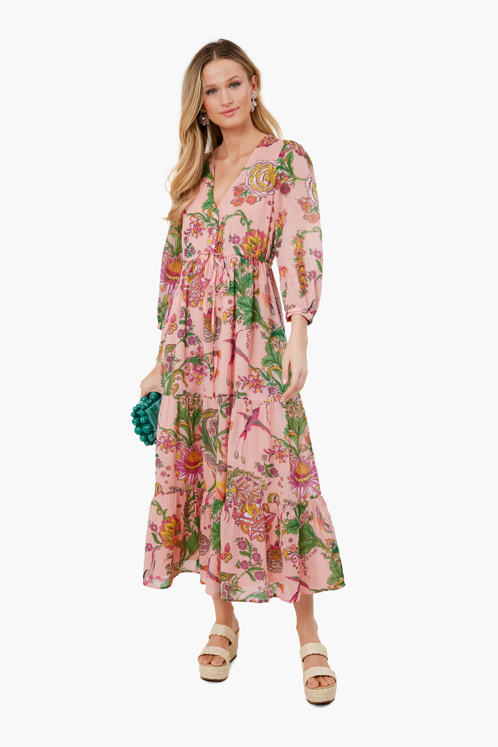 A woman wears a Banjanan organic cotton pink floral midi dress with three-quarter sleeves, holds a small teal clutch, and pairs the look with beige sandals.