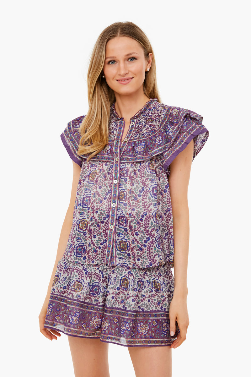 A woman wears a short purple patterned dress with ruffled sleeves by Sea New York, styled like a flutter sleeve top.