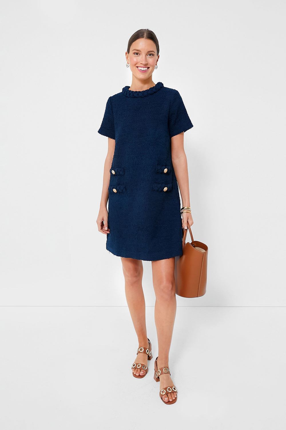 A woman wears a Tuckernuck navy tweed shift dress with vintage-inspired buttons, tan sandals, a brown bucket bag, and accessorizes with bracelets and earrings, smiling brightly.