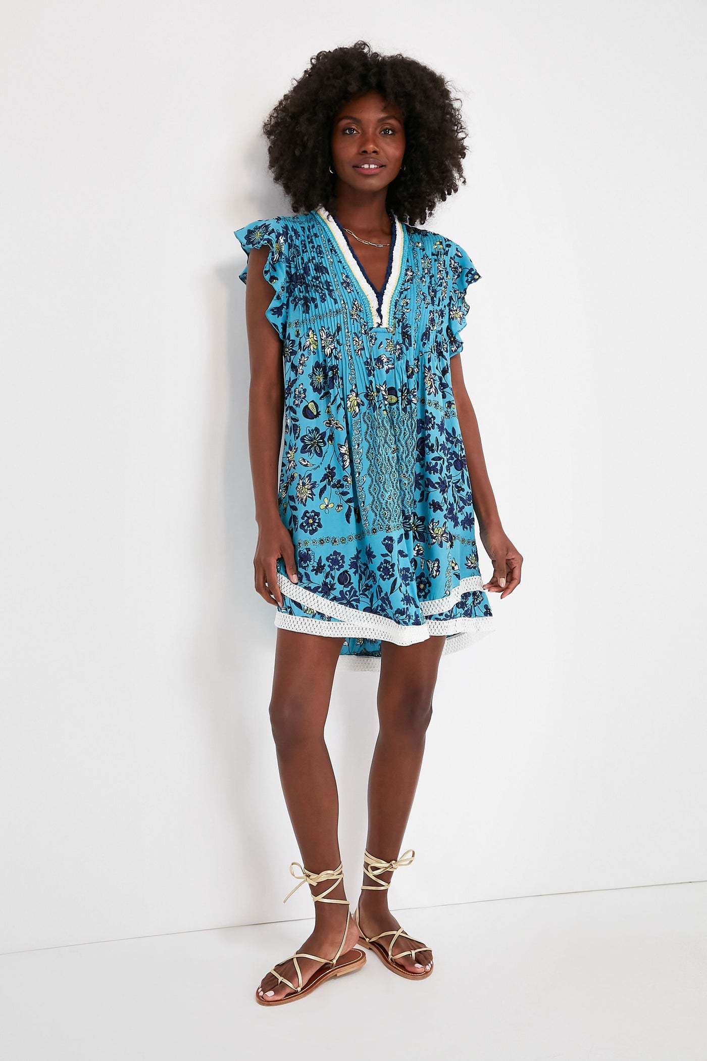 A woman wears the Poupette St Barth Sasha Dress, a short blue floral boho-chic piece with ruffled sleeves and lace trim, paired with tan lace-up sandals.