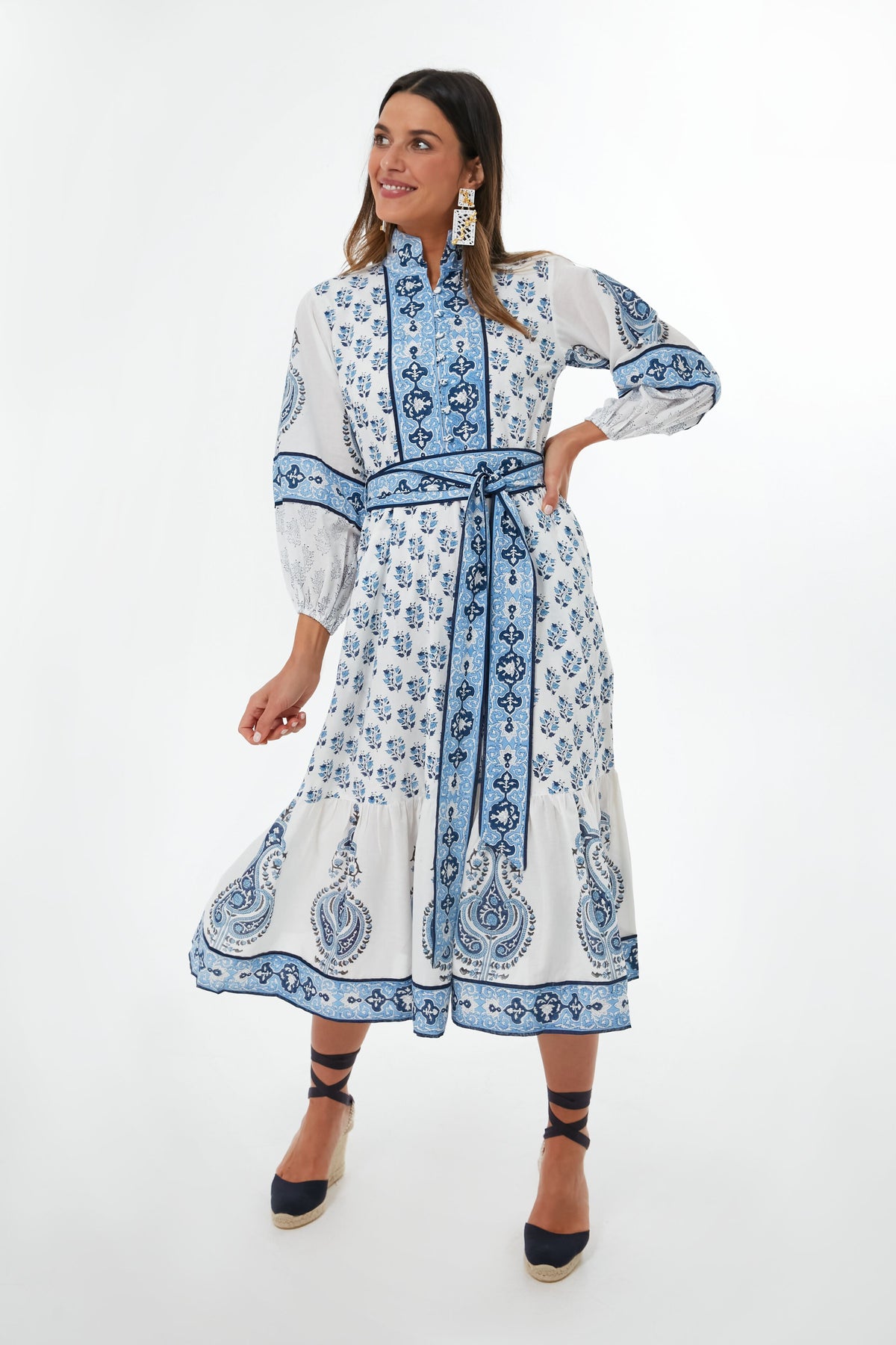 A woman wears the Sue Sartor Sky Blue Cornflower Flounce Dress—a white and blue patterned, long sleeve midi caftan crafted from sustainable fabrics—styled with a matching belt, black espadrille wedges, and statement earrings.