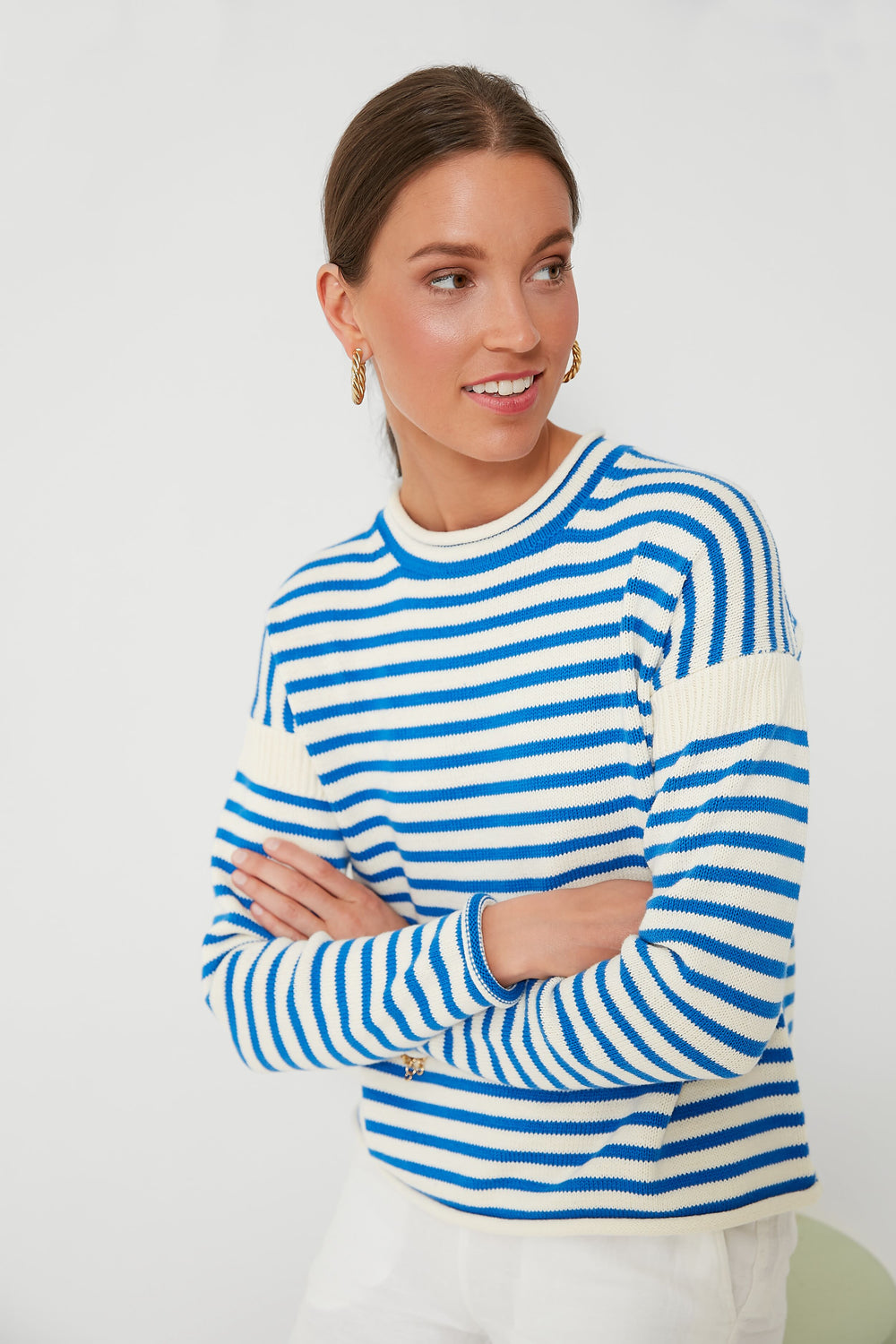 A woman in an Alex Mill blue and white mariner stripe sweater with gold hoop earrings stands with arms crossed, looking to the side.