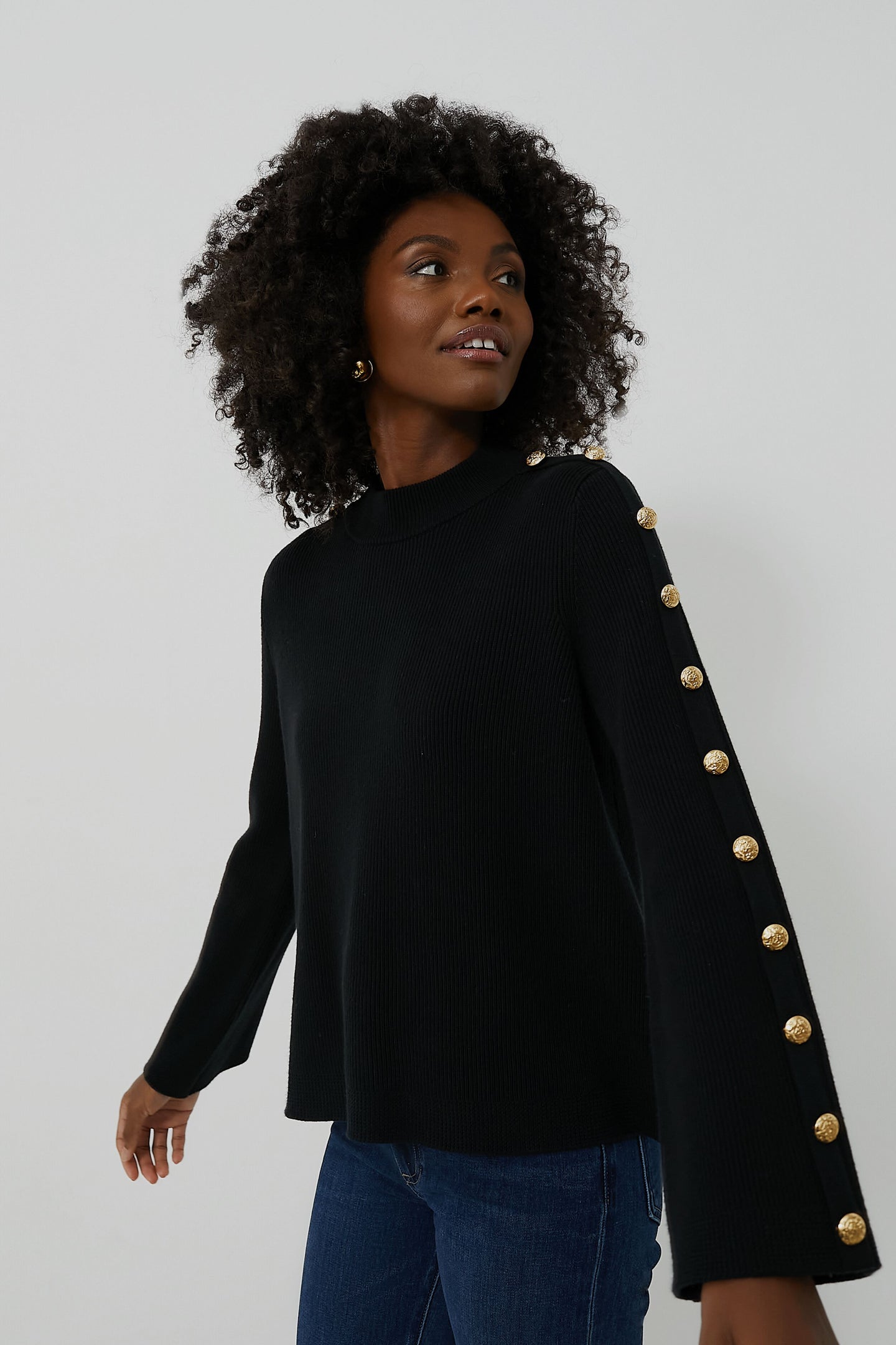 A woman wears a Tuckernuck Monroe Sweater, a black ribbed knit with gold button accents on the sleeves, paired with blue jeans.