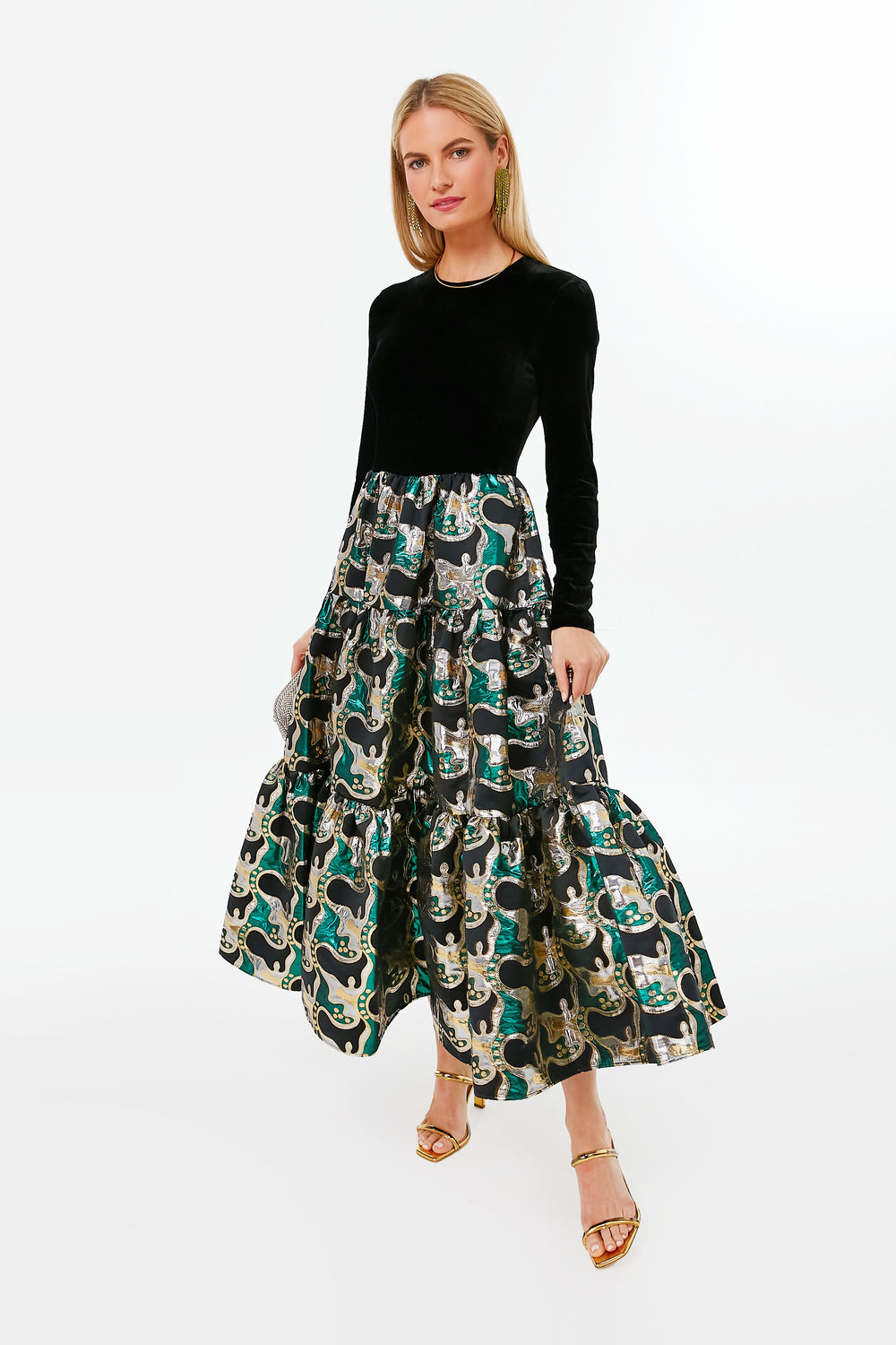A woman stands poised in elegant fashion, wearing a long black velvet La DoubleJ evening dress with a metallic patterned skirt and gold sandals.