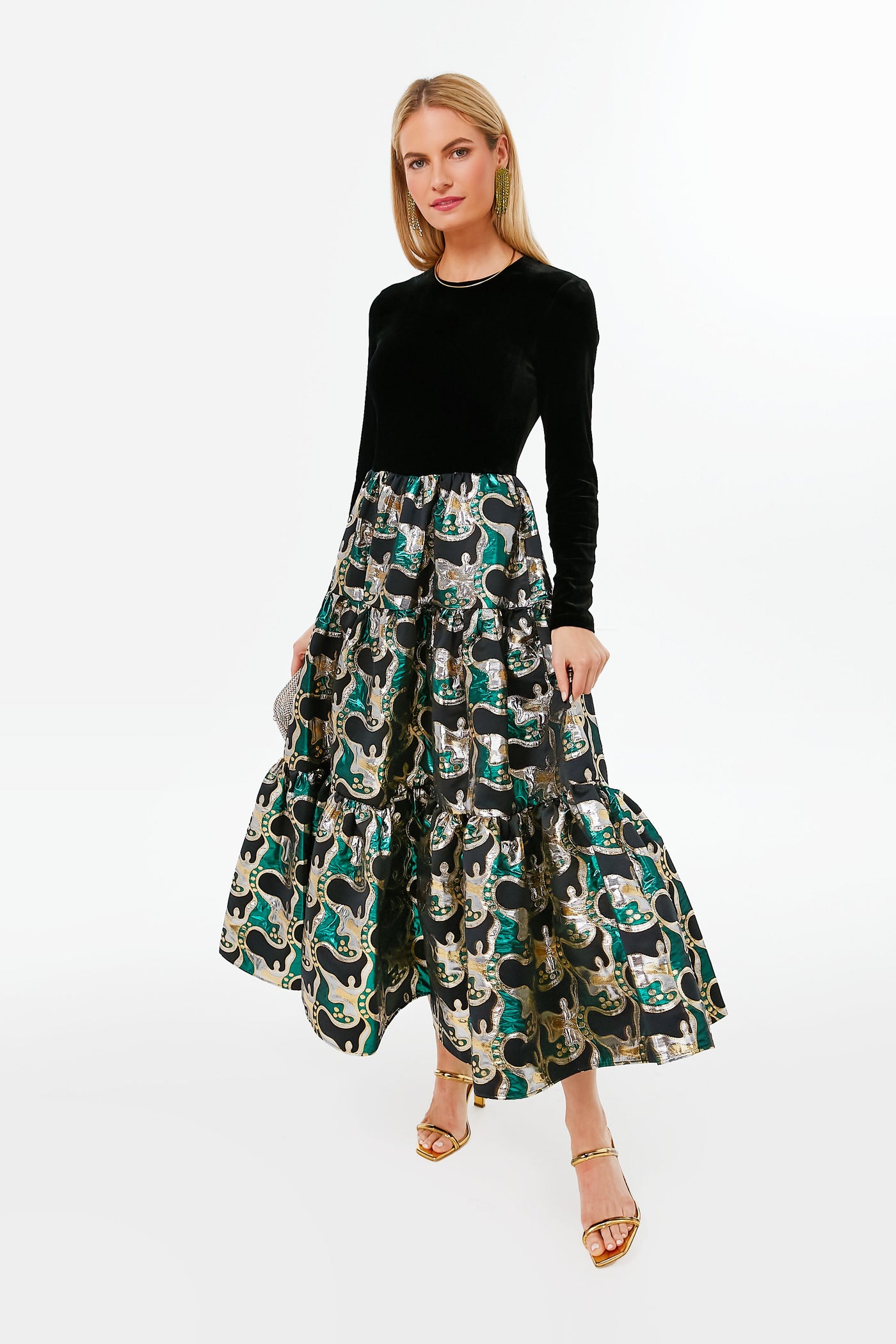 A woman stands poised in elegant fashion, wearing a long black velvet La DoubleJ evening dress with a metallic patterned skirt and gold sandals.