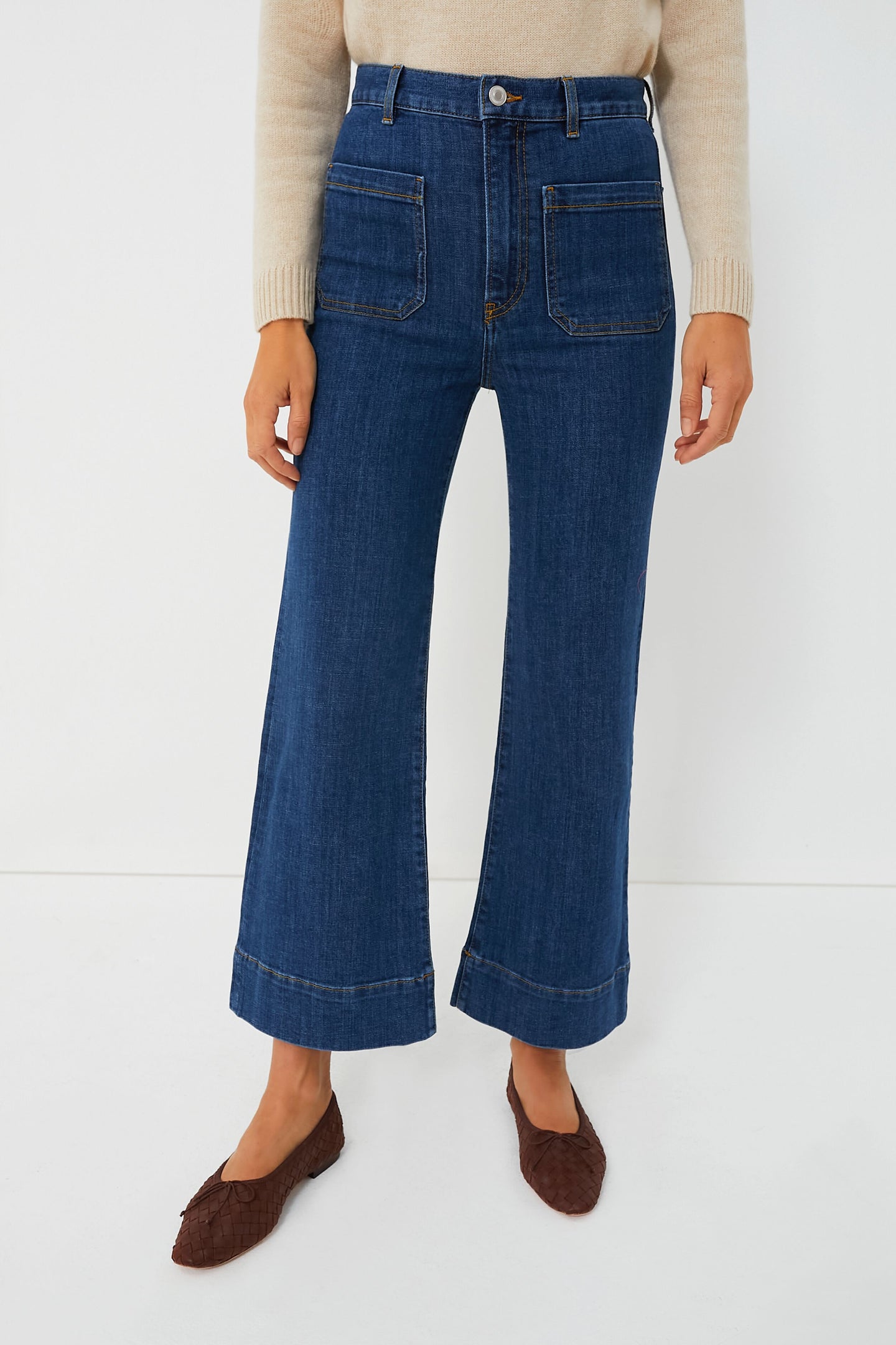 A person wears Jeanerica high-waisted, wide-leg organic denim jeans with front patch pockets, paired with a beige sweater and brown woven flats.