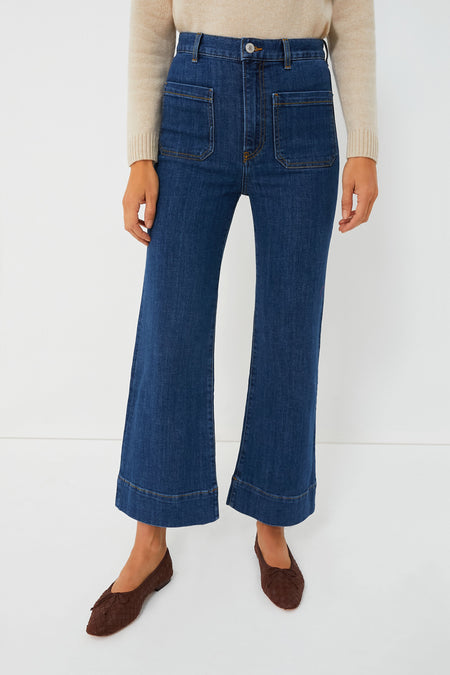 A person wears Jeanerica high-waisted, wide-leg organic denim jeans with front patch pockets, paired with a beige sweater and brown woven flats.