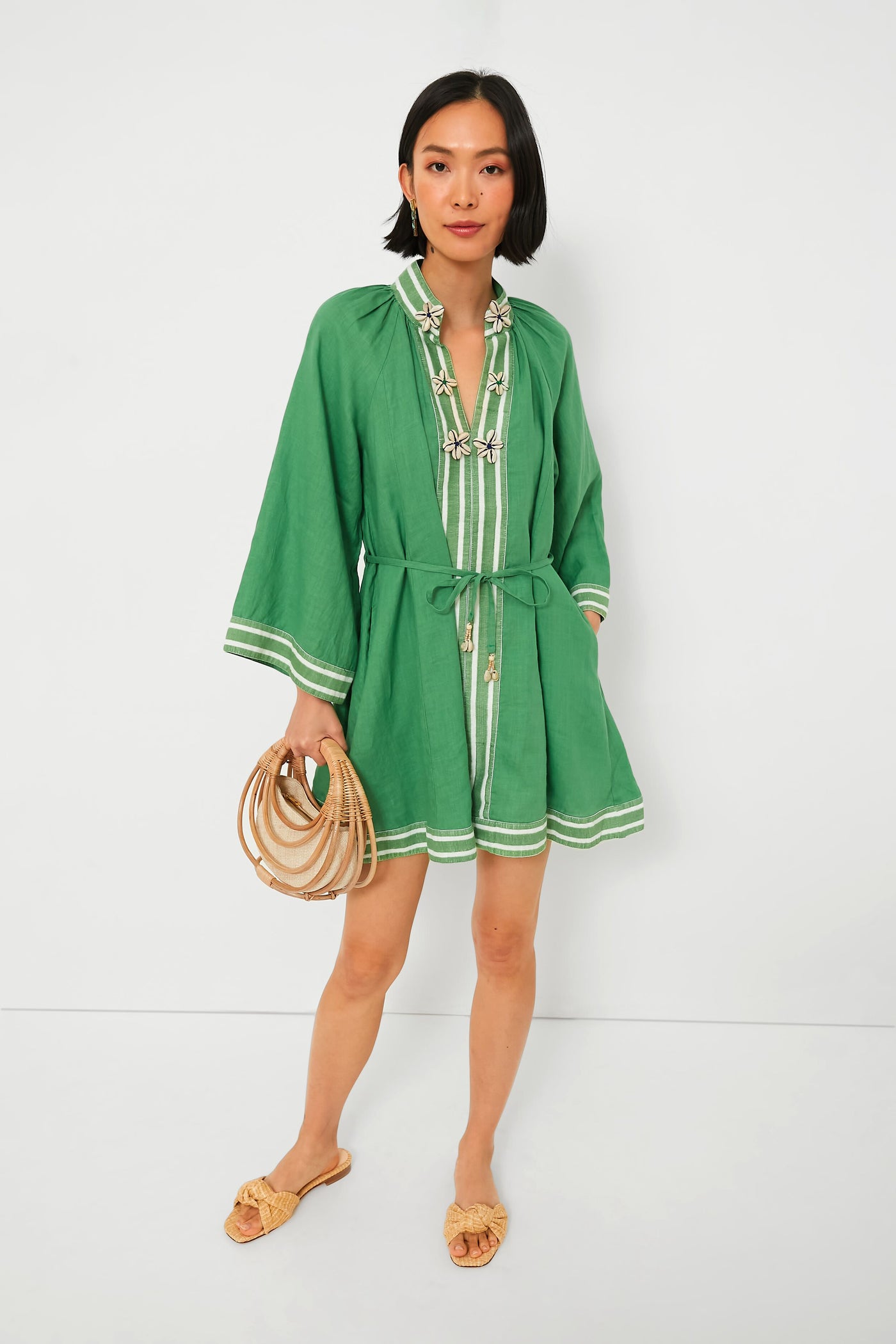 A woman showcases the Hannah Artwear Arlo Lumi Dress, a green belted linen dress with white trim, styled with a round straw bag and tan flat sandals—an elegant womenswear choice.
