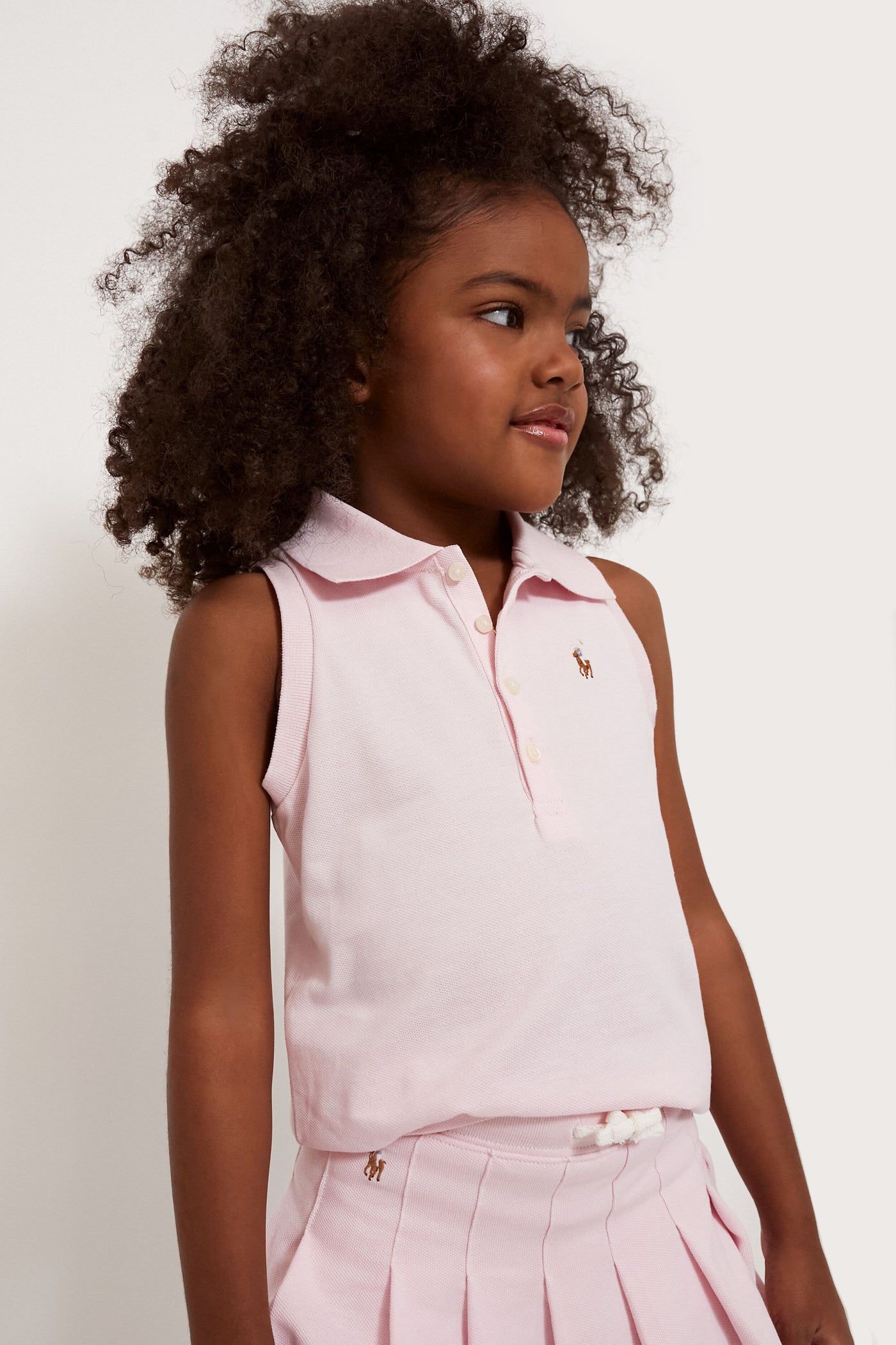 Young girl with curly hair wearing a sleeveless light pink Polo Ralph Lauren shirt and matching pleated skirt.