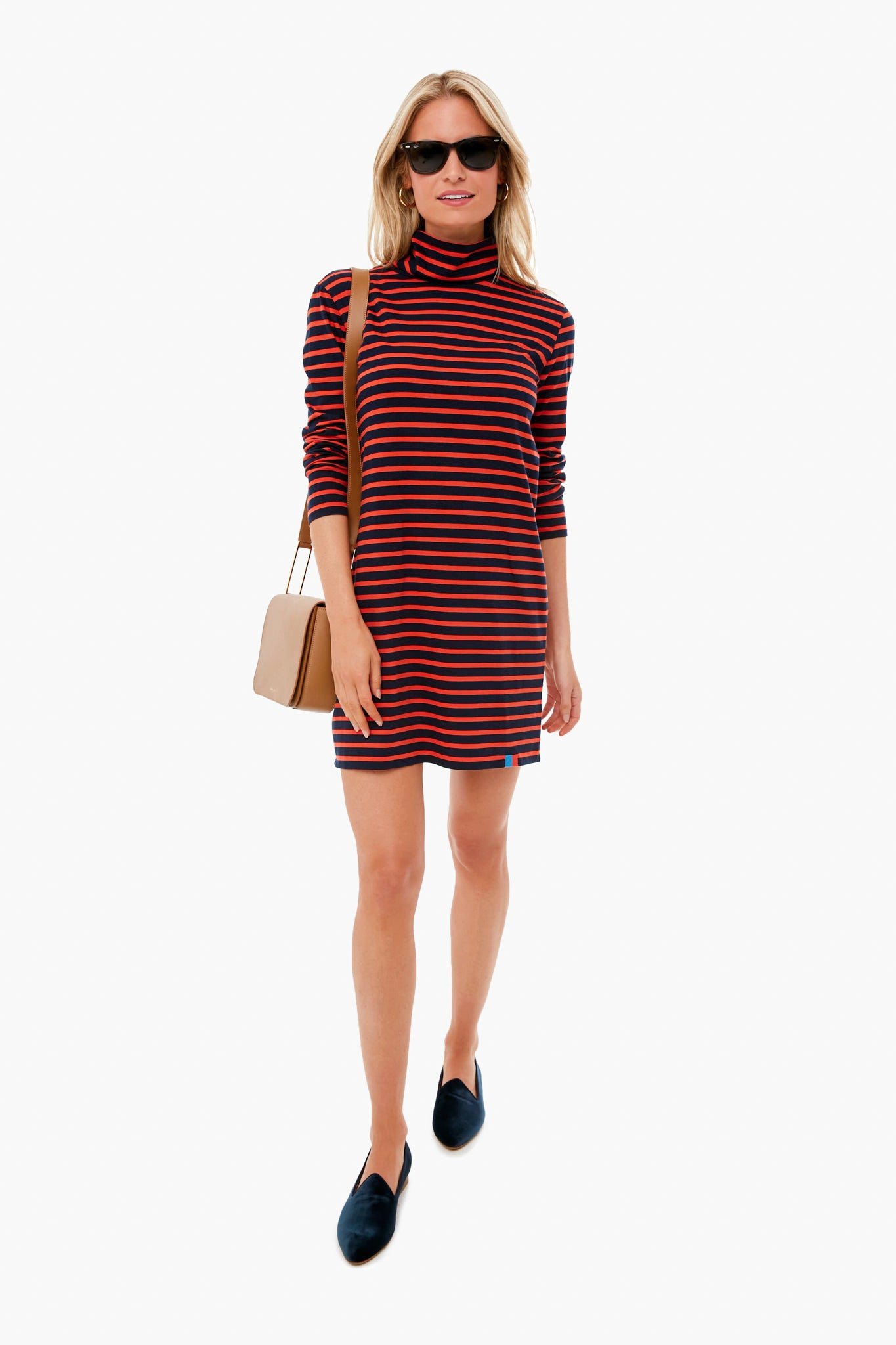 A woman wearing sunglasses, a KULE striped turtleneck dress, black shoes, and a beige shoulder bag stands confidently.