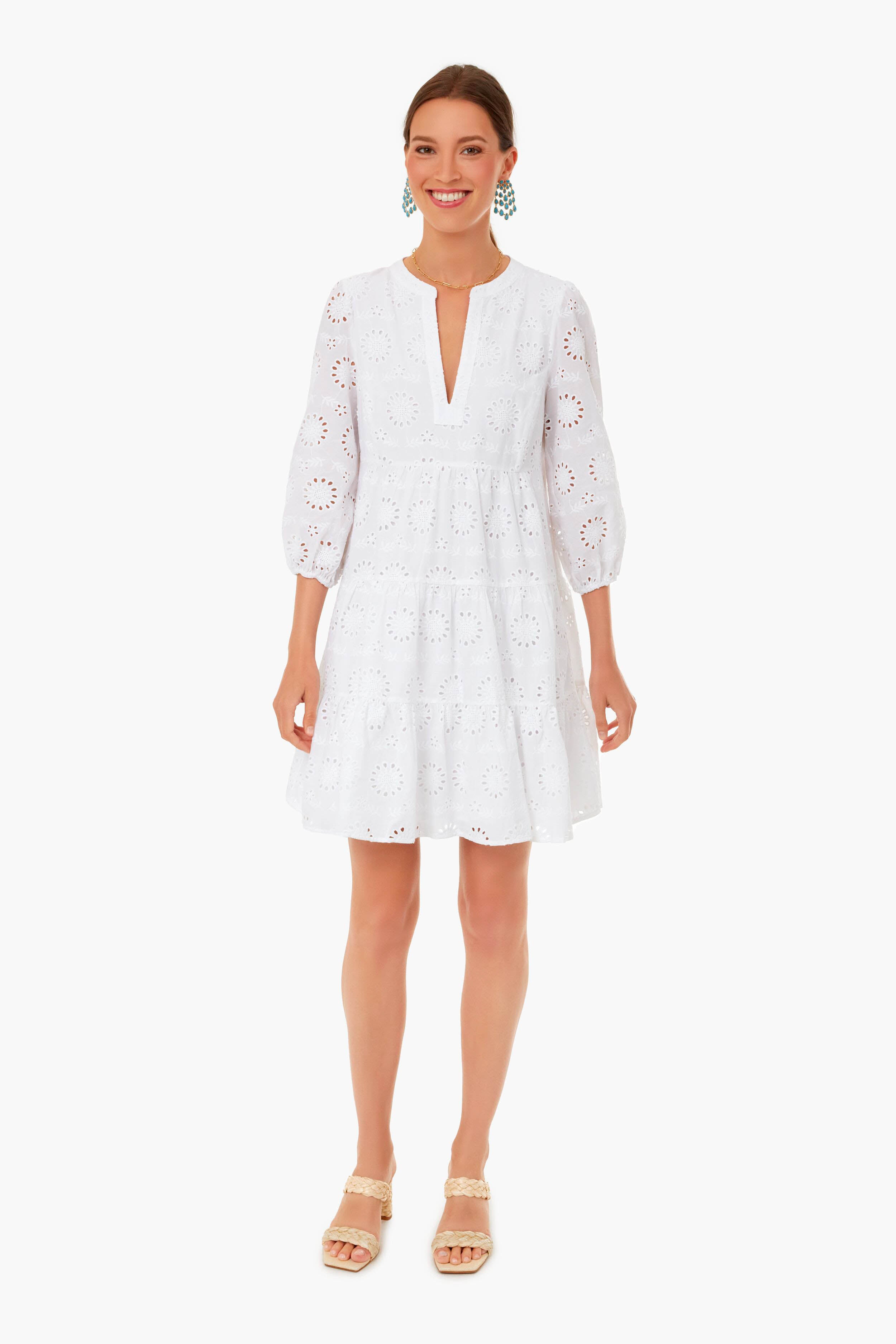 A woman stands facing forward, smiling in a white, knee-length eyelet tunic dress with three-quarter sleeves by Sail to Sable and beige wedge sandals—an effortlessly chic choice for women’s resort wear. Her hair is pulled back.