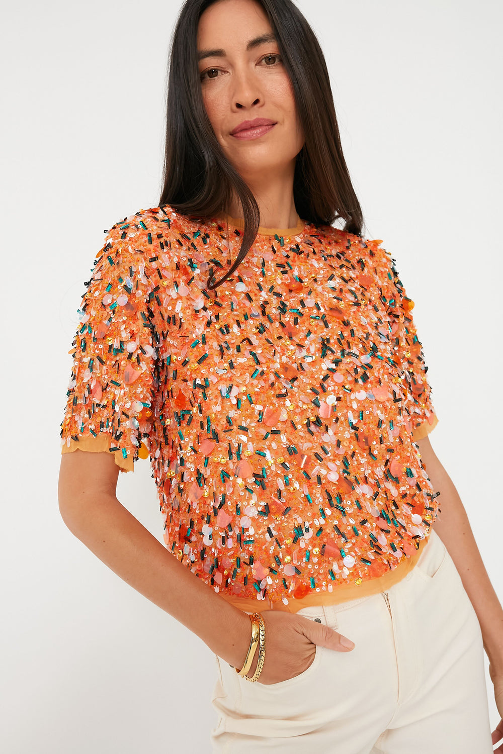 A woman wears a Hunter Bell Orange Shelby Top with padded shoulders and multicolored sequin embellishments, paired with cream pants.