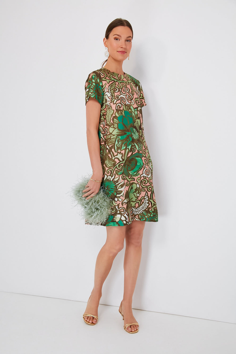 A woman wears a short-sleeved green and beige patterned silk twill dress from Milanese brand La DoubleJ, tan heels, and holds a pale green feathered clutch.