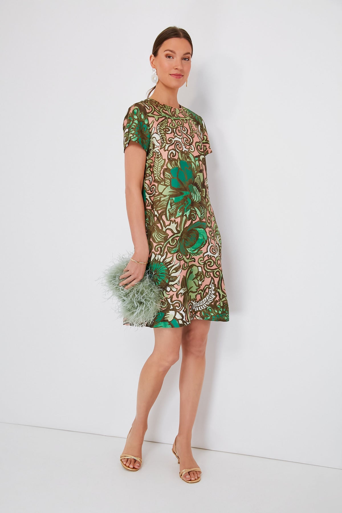 A woman wears a short-sleeved green and beige patterned silk twill dress from Milanese brand La DoubleJ, tan heels, and holds a pale green feathered clutch.