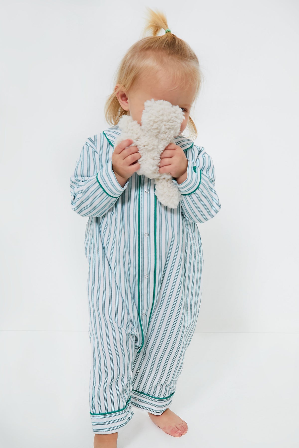 A small child in striped pajamas from Petite Plume stands barefoot, holding a plush toy to their face while wearing an emerald ticking romper.