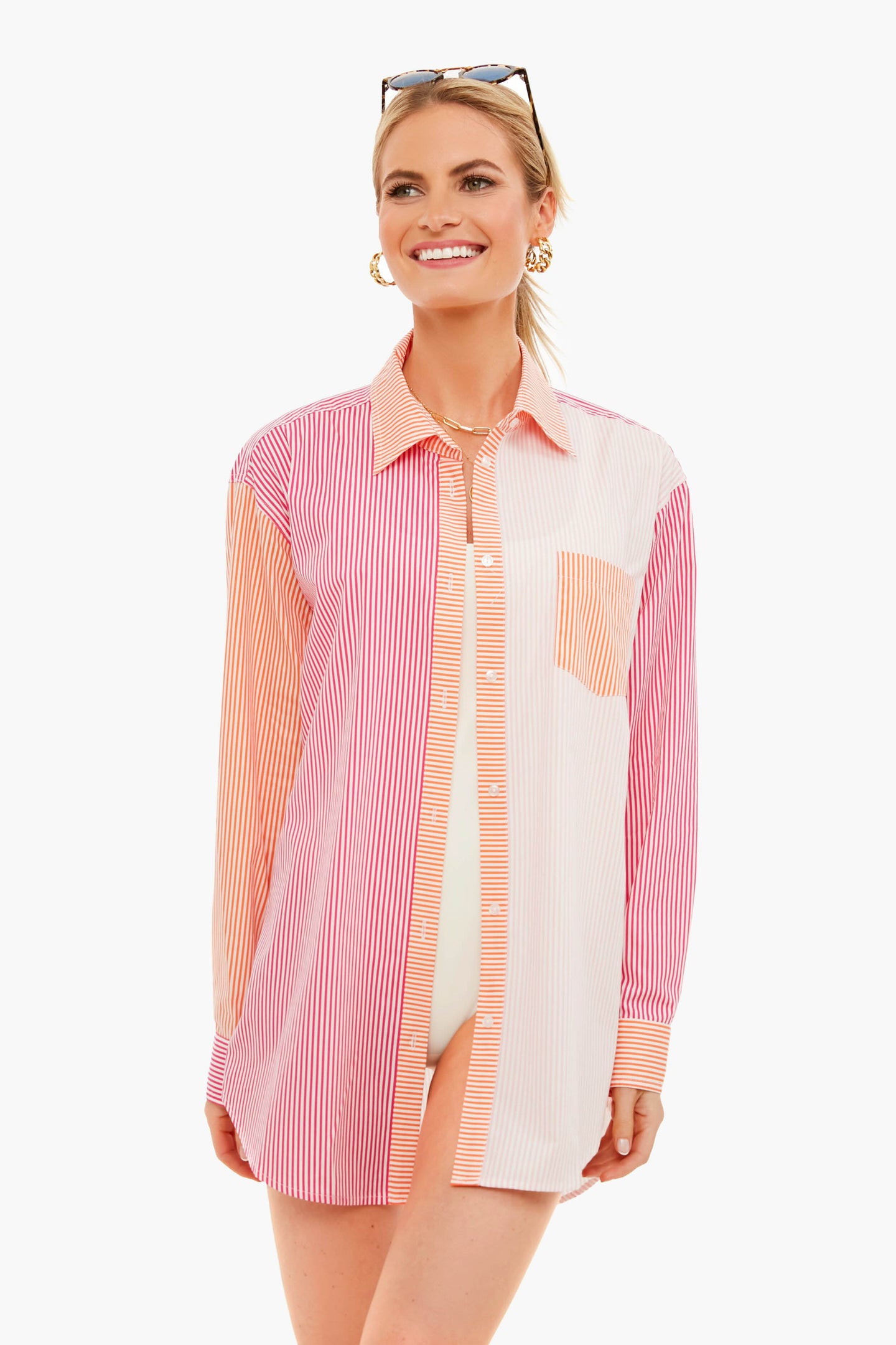 A woman wearing a Solid & Striped long-sleeve, multicolored striped oxford tunic with gold hoop earrings and sunglasses on her head stands smiling.