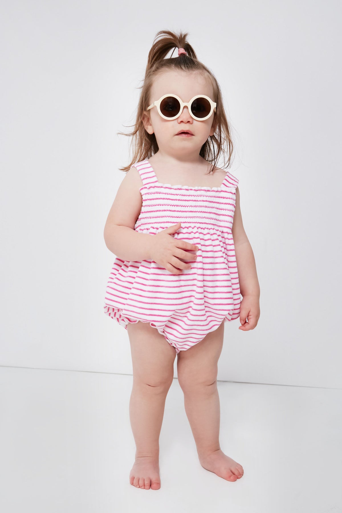A toddler in kissy kissy round sunglasses and a pink-and-white striped smocked bubble romper stands barefoot, showing off cozy Pima cotton baby clothing.