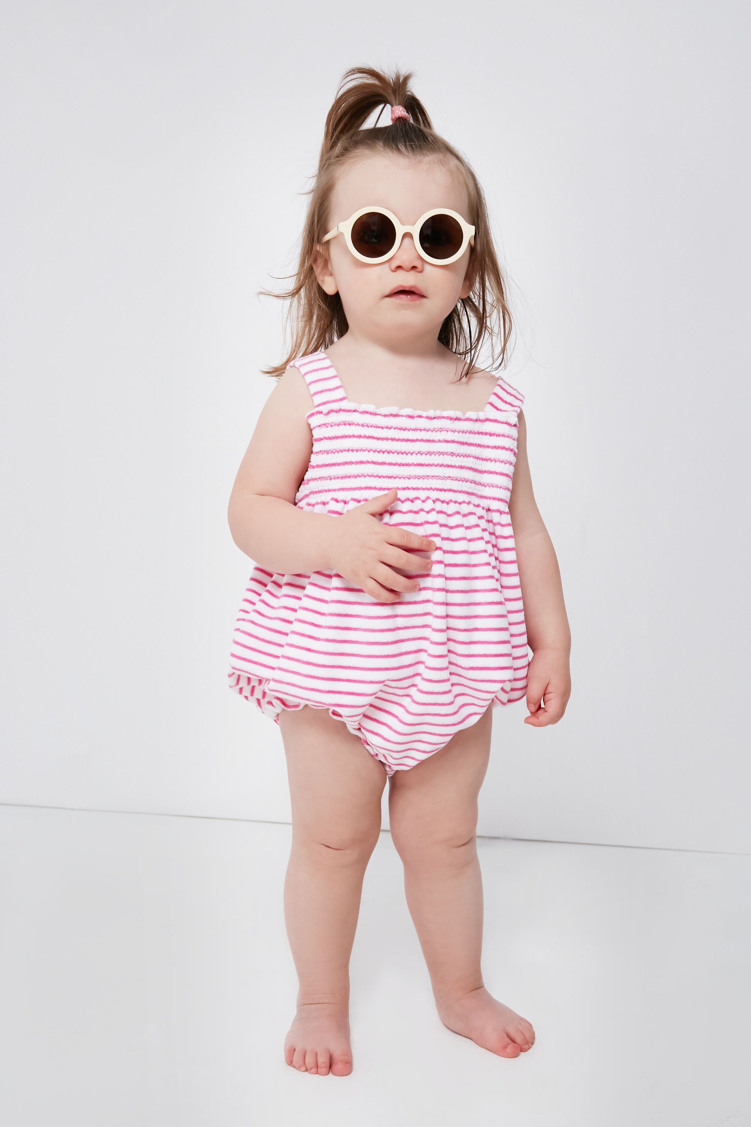 A toddler in kissy kissy round sunglasses and a pink-and-white striped smocked bubble romper stands barefoot, showing off cozy Pima cotton baby clothing.
