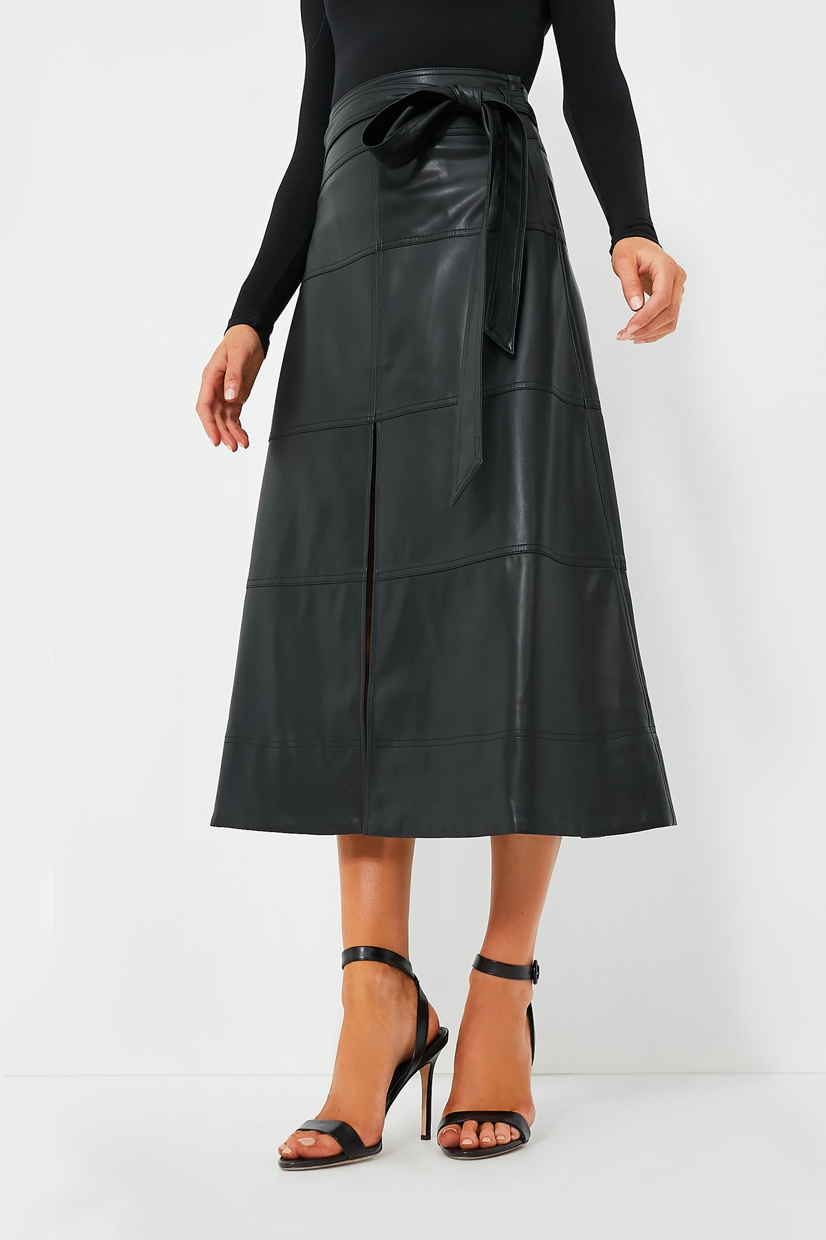 A person wears a Tanya Taylor black high-waisted vegan leather midi skirt with a front slit and tie belt, styled with a black long-sleeve top and black heeled sandals.