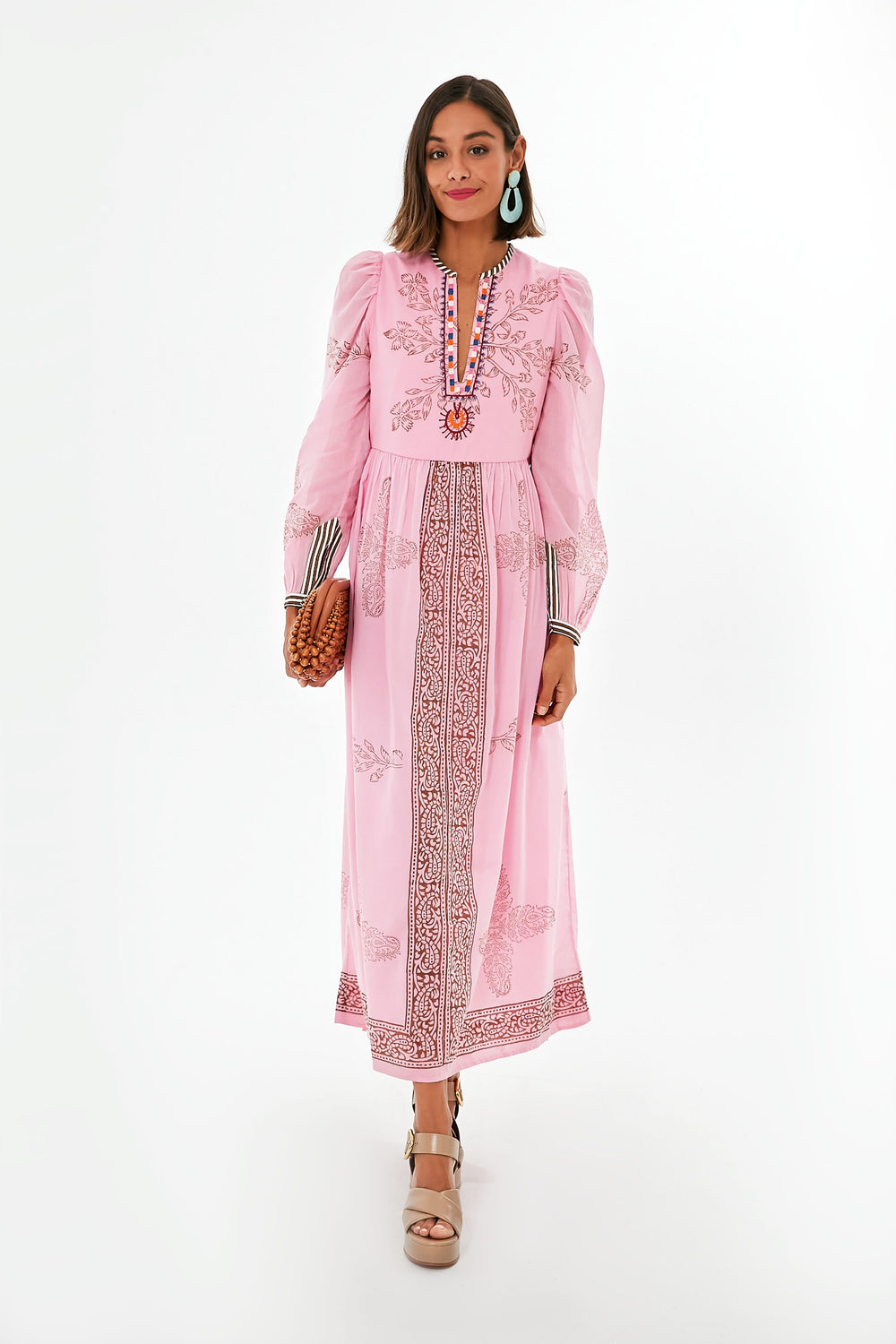 A woman in an Alix of Bohemia long pink hand block printed cotton boho maxi dress with floral patterns, statement earrings, platform sandals, and a brown woven clutch.