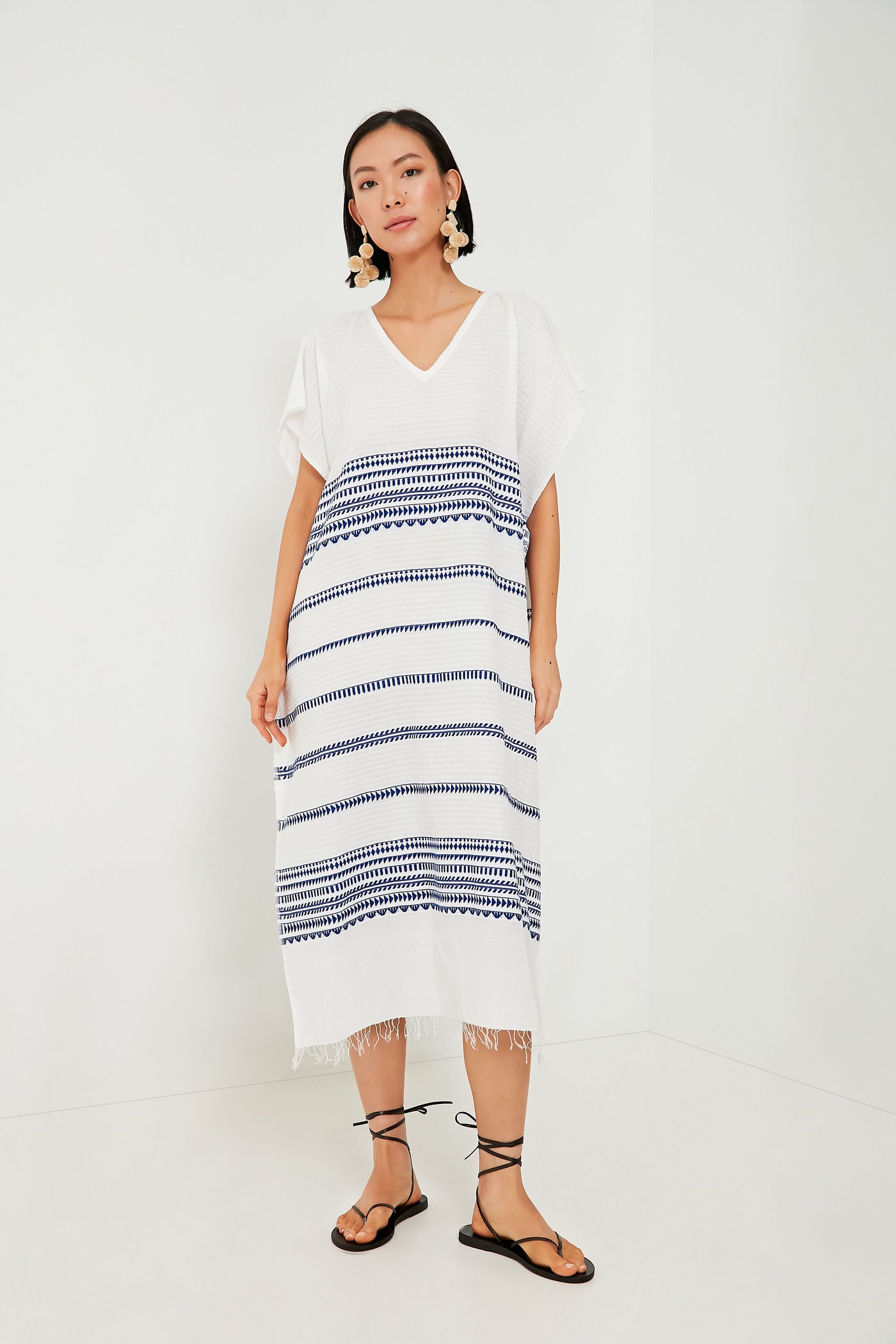A woman stands in a white room wearing a lemlem handmade midi dress featuring blue horizontal patterns, paired with black sandals and large statement earrings, evoking the style of an Ethiopian caftan.