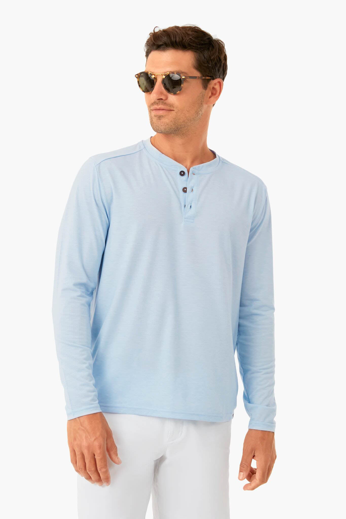 A man confidently stands wearing Fair Harbor sunglasses and a light blue UPF 50 Henley shirt, crafted from recycled plastic bottles, paired with white pants.