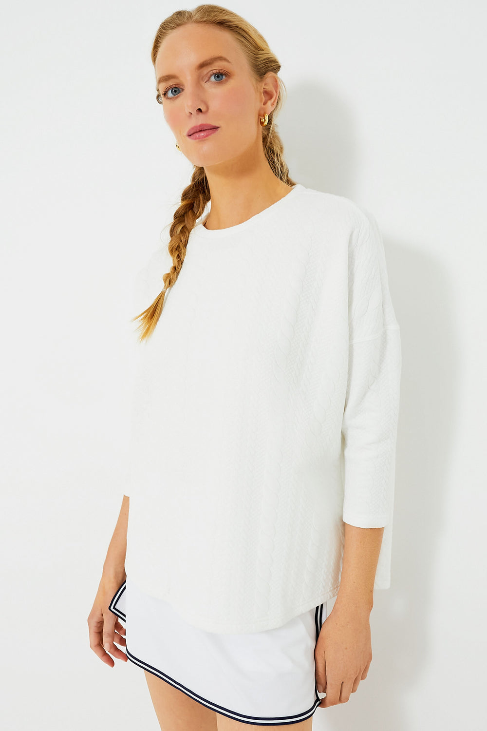 A woman with a braided hairstyle models Tnuck Sport’s white quilted long-sleeve top and matching white skirt with black trim, both featuring a relaxed fit.