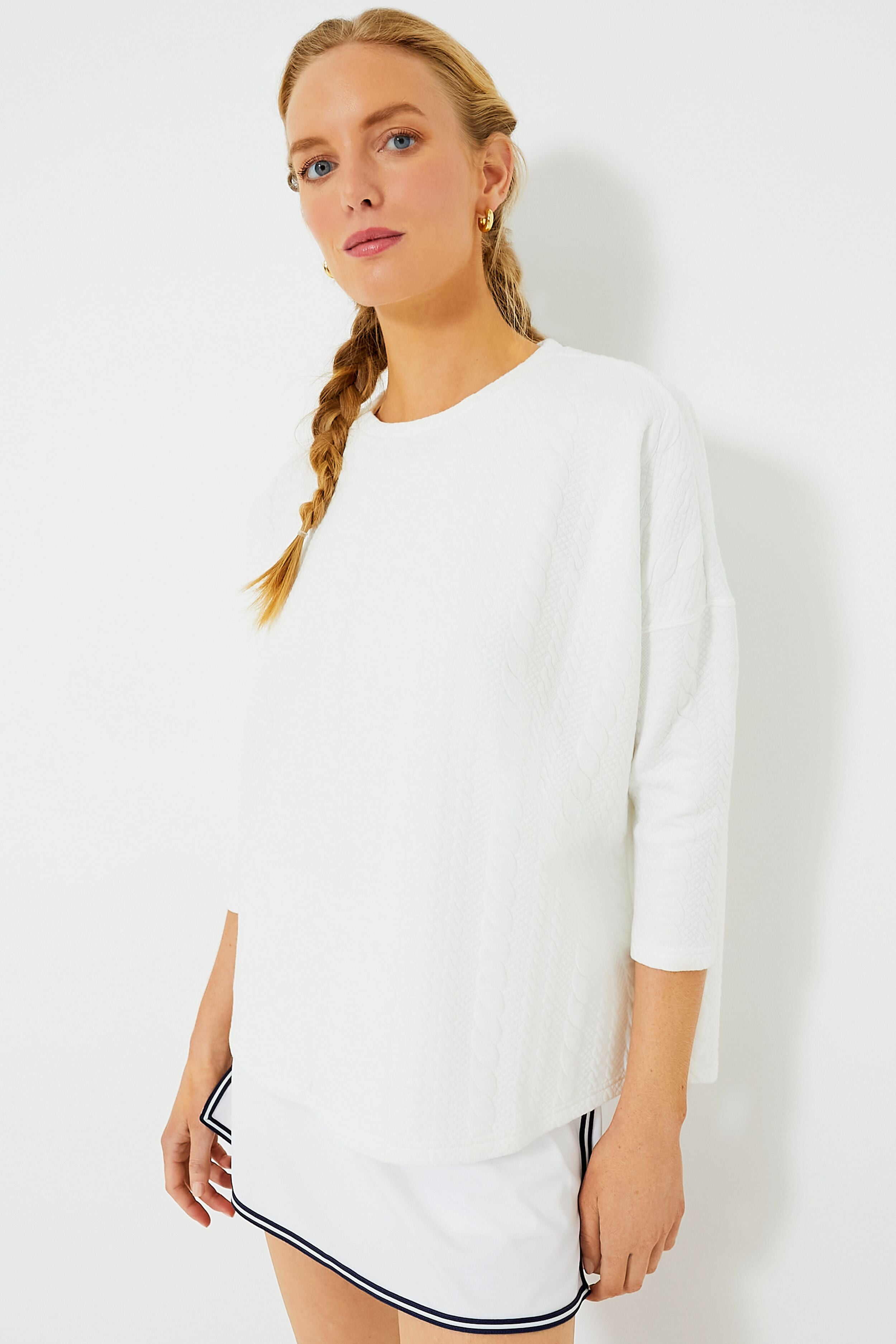 A woman with a braided hairstyle models Tnuck Sport’s white quilted long-sleeve top and matching white skirt with black trim, both featuring a relaxed fit.