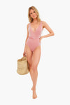 A woman stands barefoot, holding a woven straw bag and wearing an onia pink belted maillot swimsuit, perfectly showcasing minimalist swimwear style.