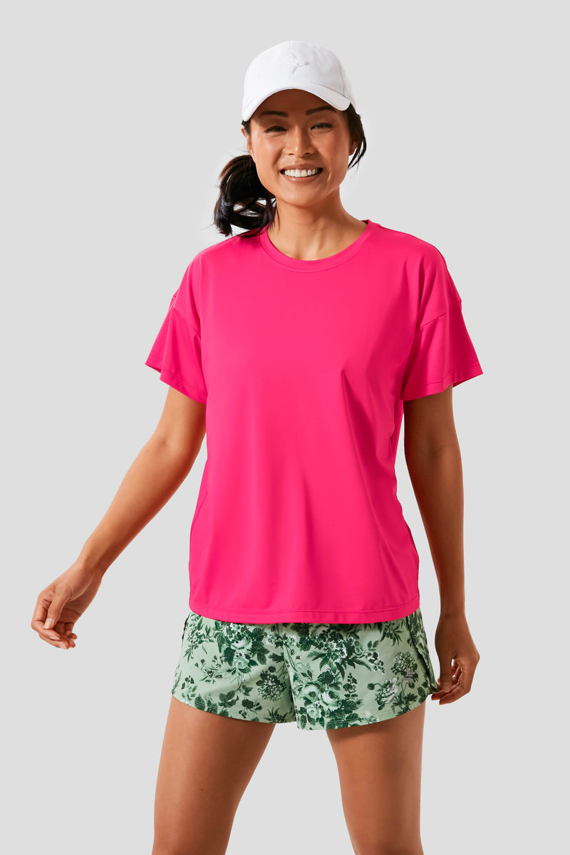 A woman smiles wearing a white cap, Tnuck Sport azalea short sleeve boyfriend tee, and green patterned shorts.