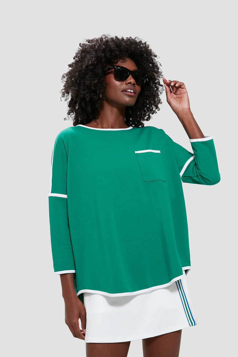 A woman wearing Tnuck Sport sunglasses, a green terry pullover with white trim and pocket, and a white skirt with blue stripes stands confidently.