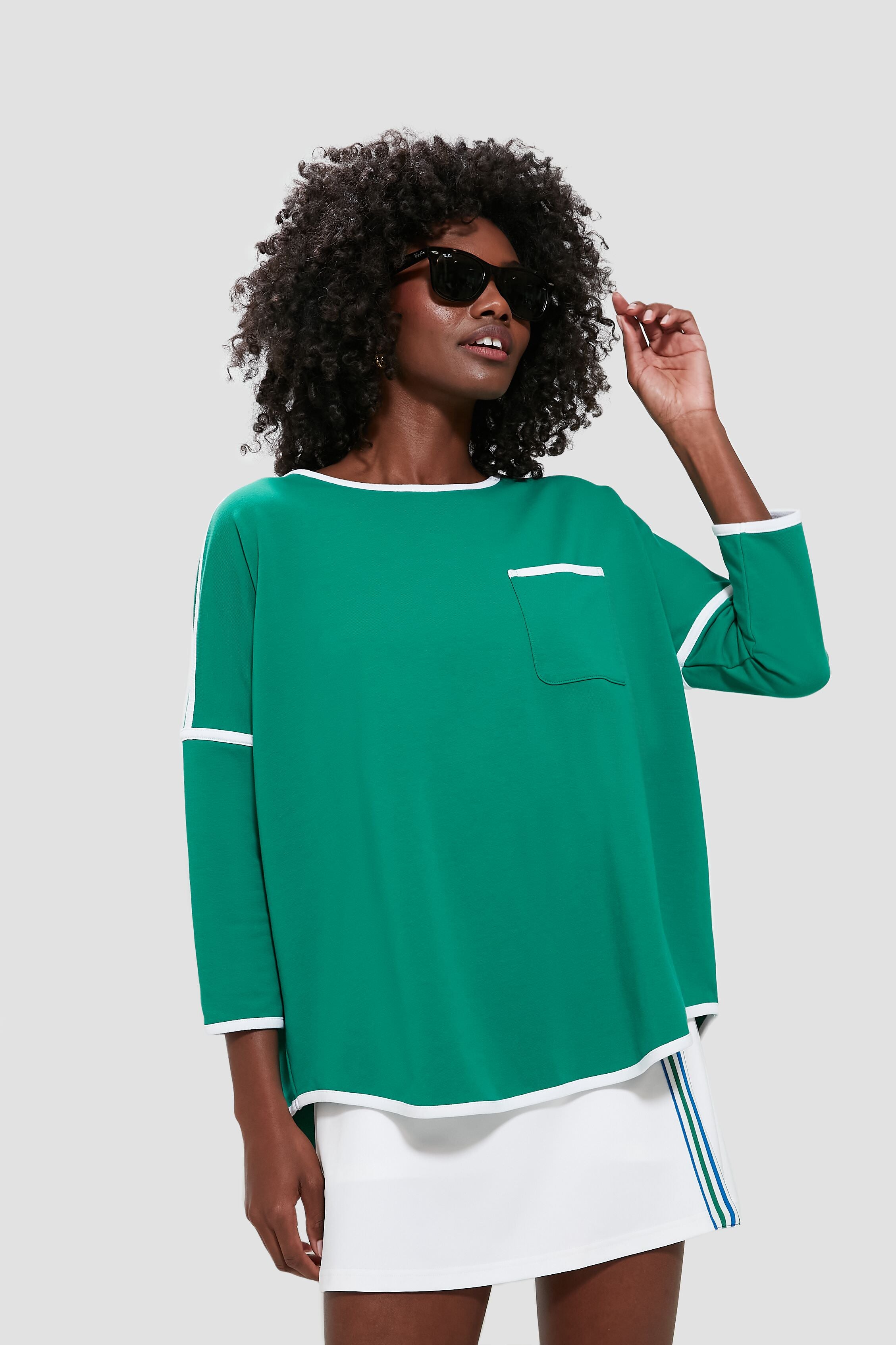 A woman wearing Tnuck Sport sunglasses, a green terry pullover with white trim and pocket, and a white skirt with blue stripes stands confidently.