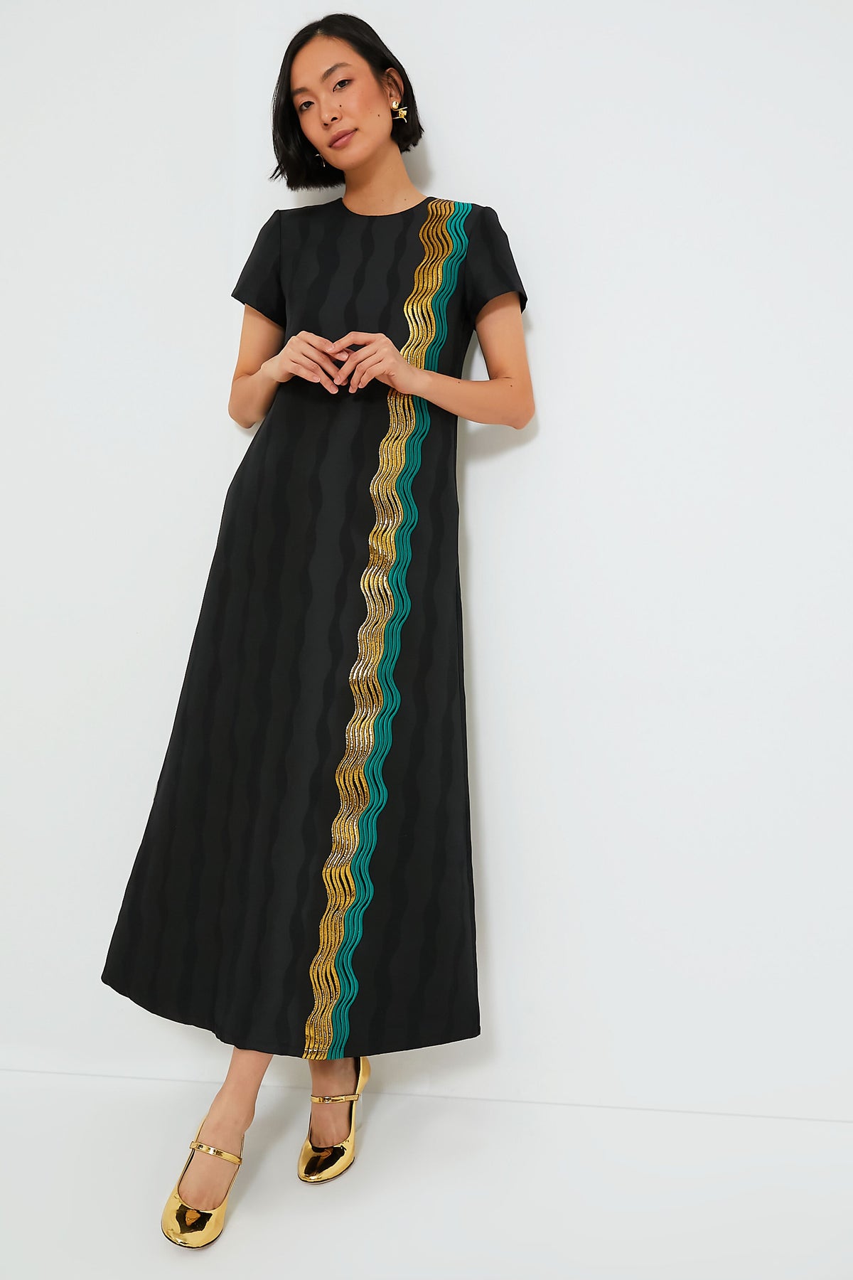 A woman wears La DoubleJ’s Jacquard The Nile Swing Dress—a black maxi accented with wavy yellow and teal stripes—paired with gold shoes.