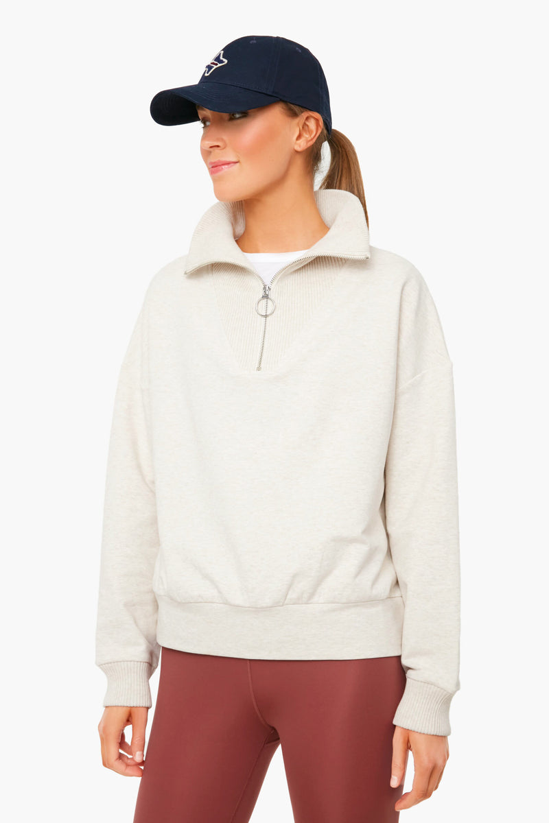 A woman wears a navy baseball cap, maroon leggings, and a beige Varley Miller Sweat pullover with a zip collar in marled cotton-blend, highlighting Varley’s relaxed-fit women’s style.