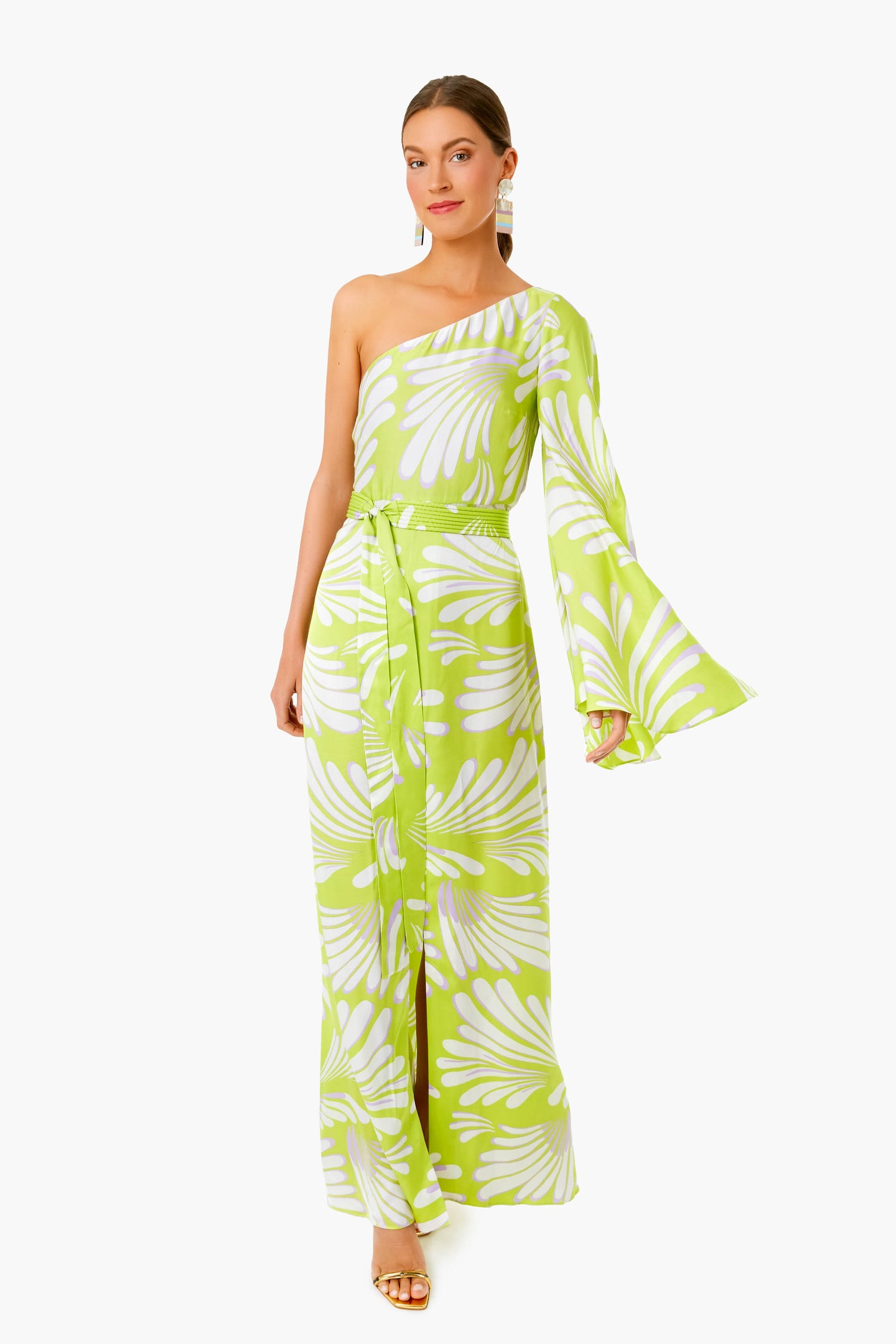 A woman wears the ALEXIS Bliss Lio Dress, a green and white one-shoulder maxi with flared sleeve and waist tie, styled with gold sandals and statement earrings for timeless womenswear elegance.