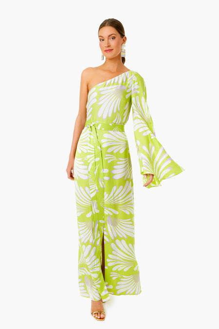 A woman wears the ALEXIS Bliss Lio Dress, a green and white one-shoulder maxi with flared sleeve and waist tie, styled with gold sandals and statement earrings for timeless womenswear elegance.