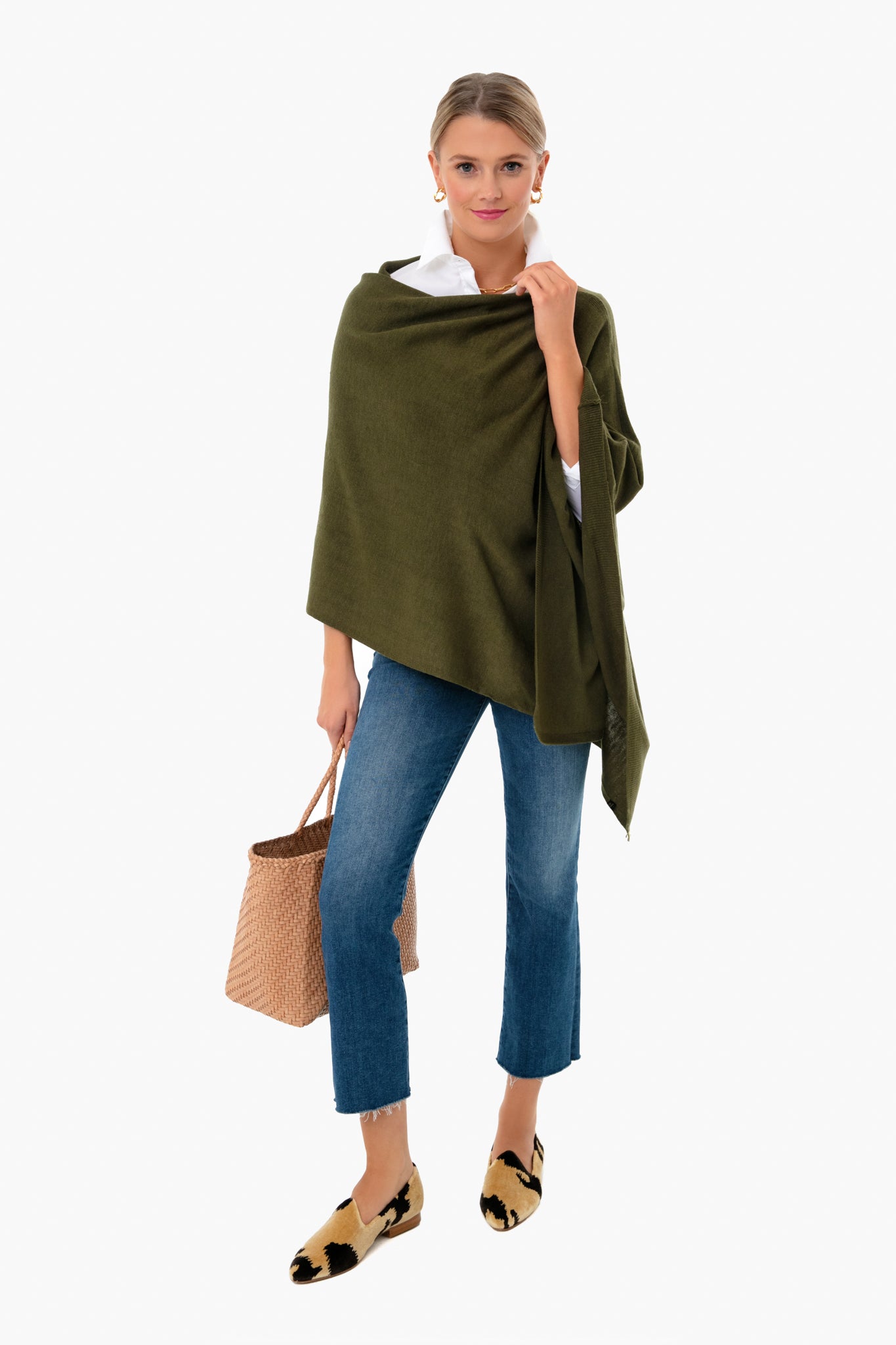 A woman stands confidently in a green breezy Poncho by Pomander Place, white shirt, blue cropped jeans, patterned loafers, and holding a woven handbag.