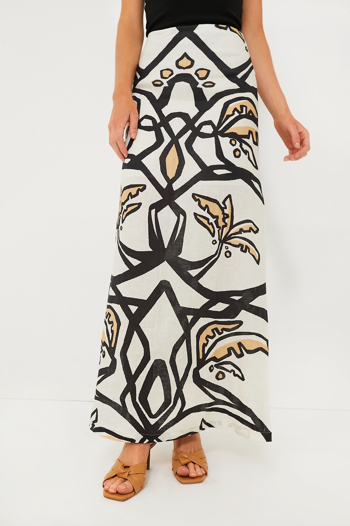 A person in an Eugenia Fernandez Malambo White Carmenza Skirt—a cream geometric print midi with bold black and tan patterns—styled with a black top and tan sandals for a sustainable fashion look.