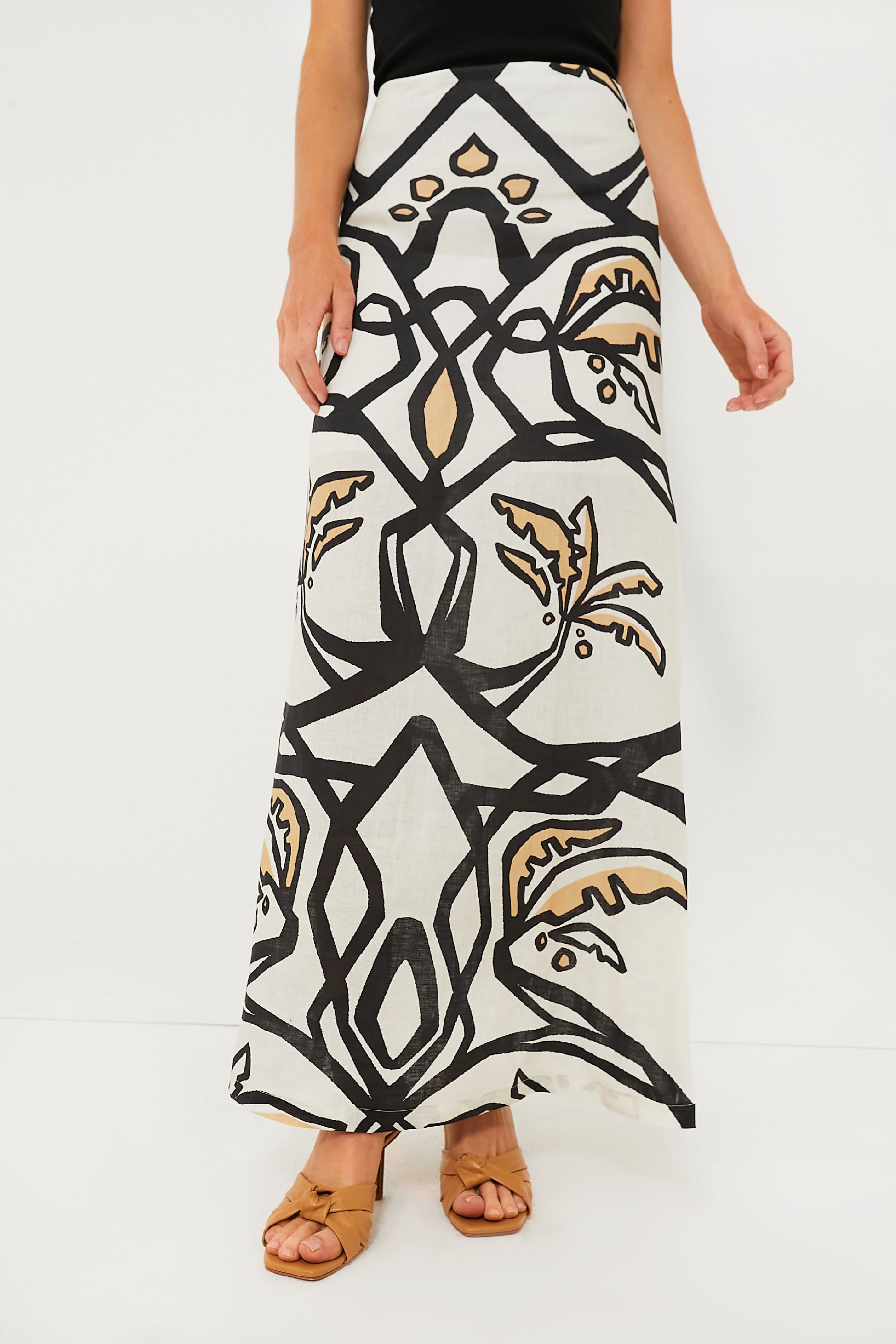 A person in an Eugenia Fernandez Malambo White Carmenza Skirt—a cream geometric print midi with bold black and tan patterns—styled with a black top and tan sandals for a sustainable fashion look.