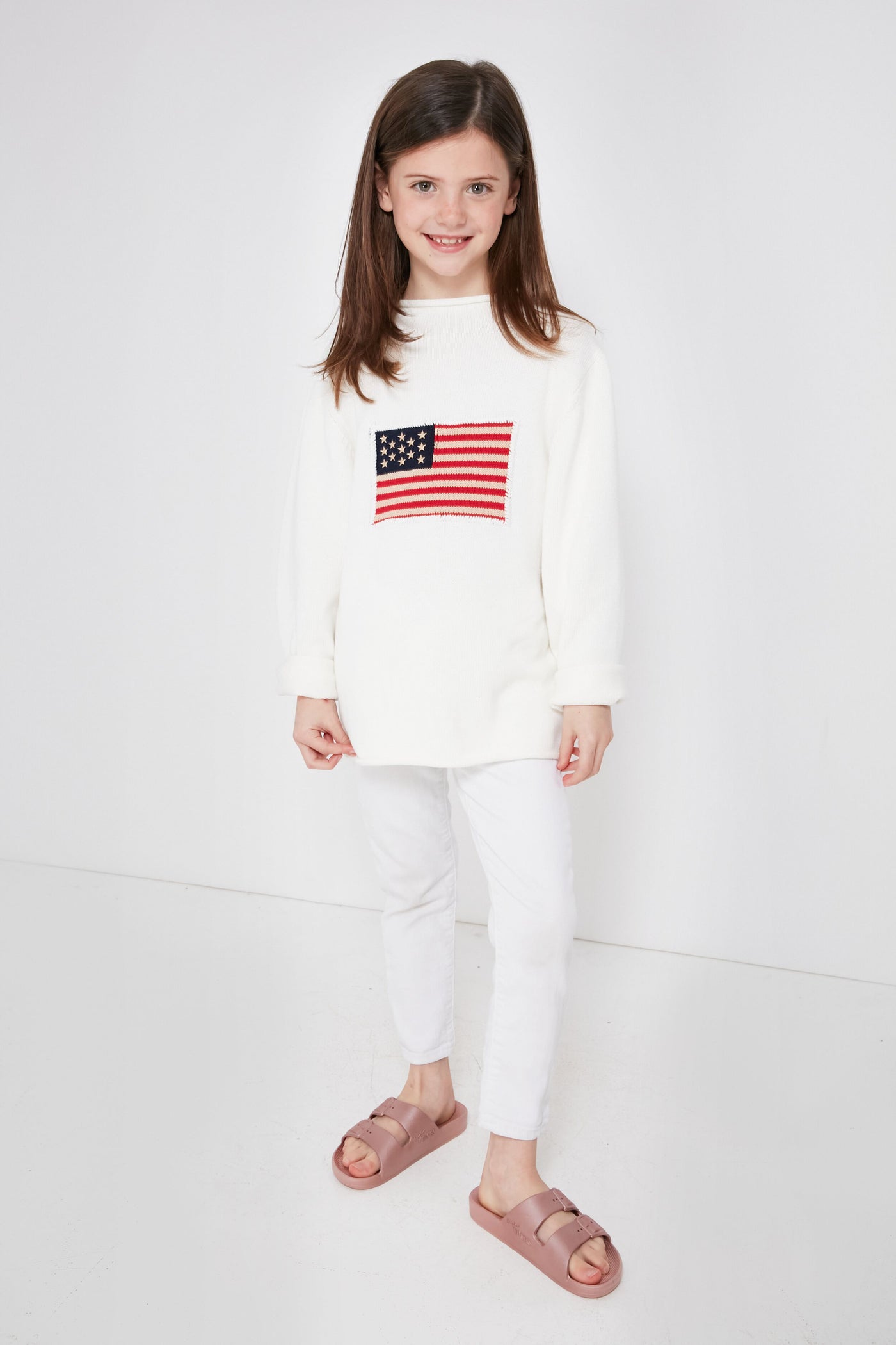 A young girl smiles at the camera wearing a Tuckernuck Tots kids flag sweater with an American flag design, paired with white pants and pink sandals.