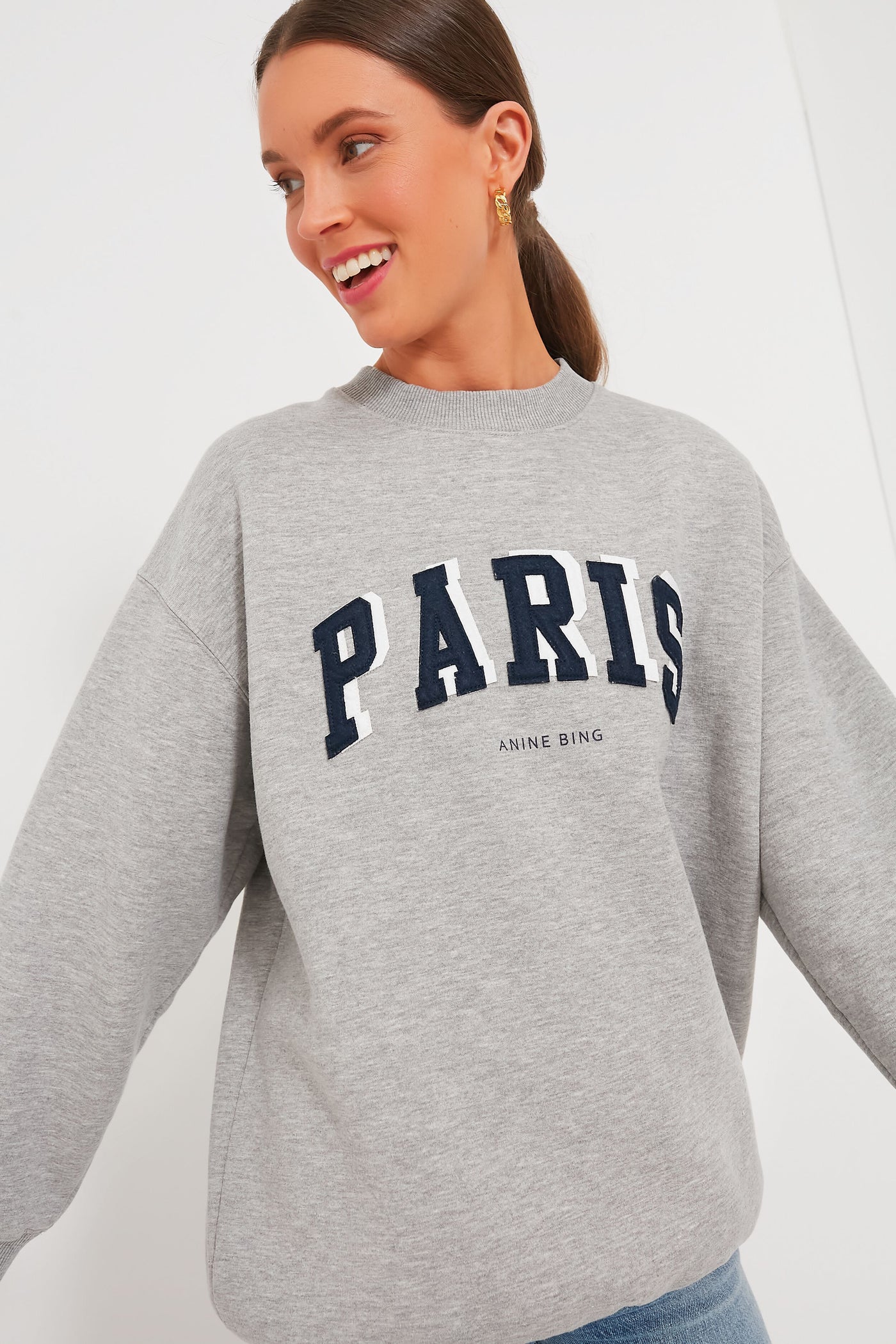A woman wears an Anine Bing oversized Paris Tyler Sweatshirt, showcasing bold black "PARIS" lettering with "ANINE BING" in smaller text below.