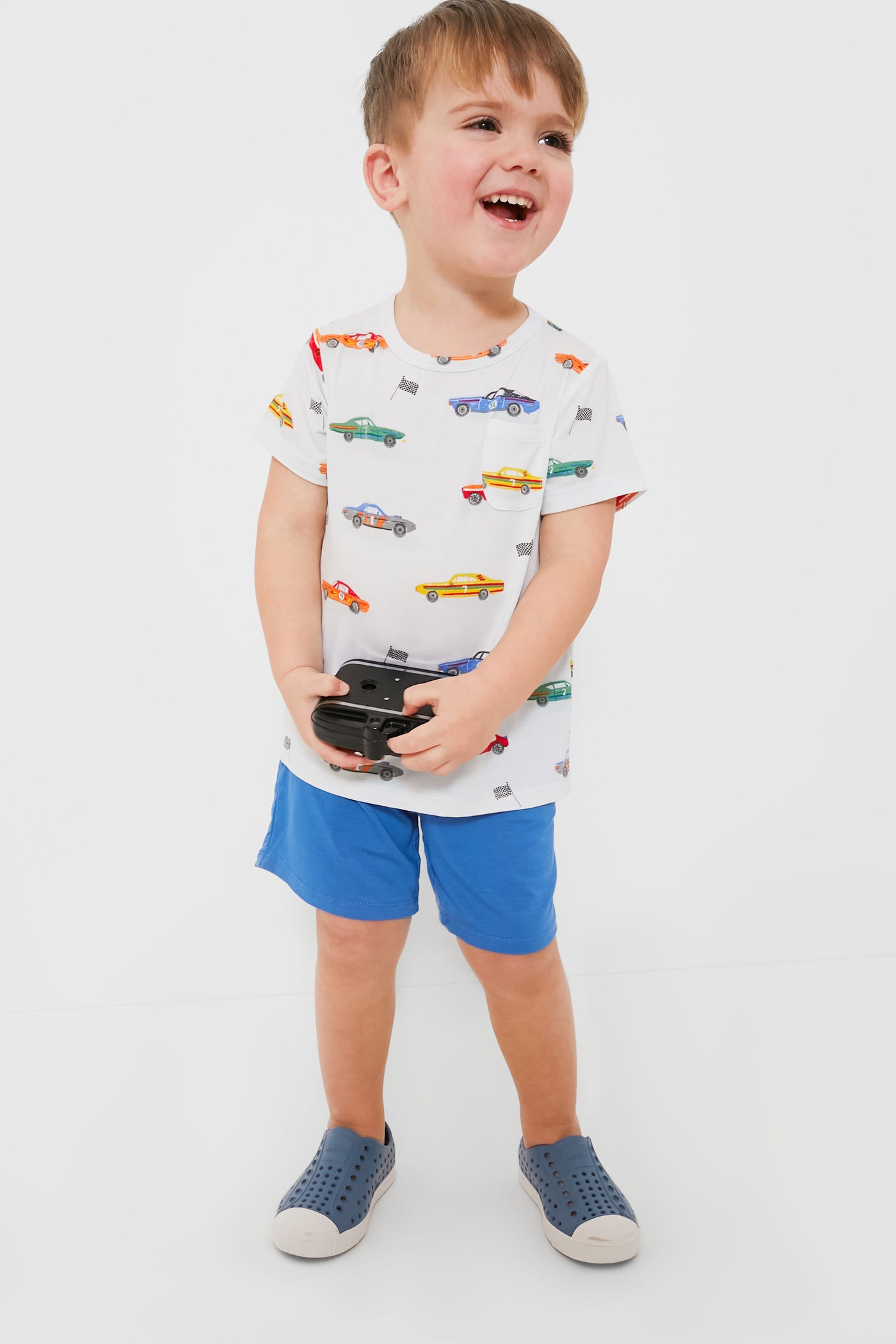 A young boy smiles while holding a black game controller, dressed in an Angel Dear muscle cars crew neck tee and short set.