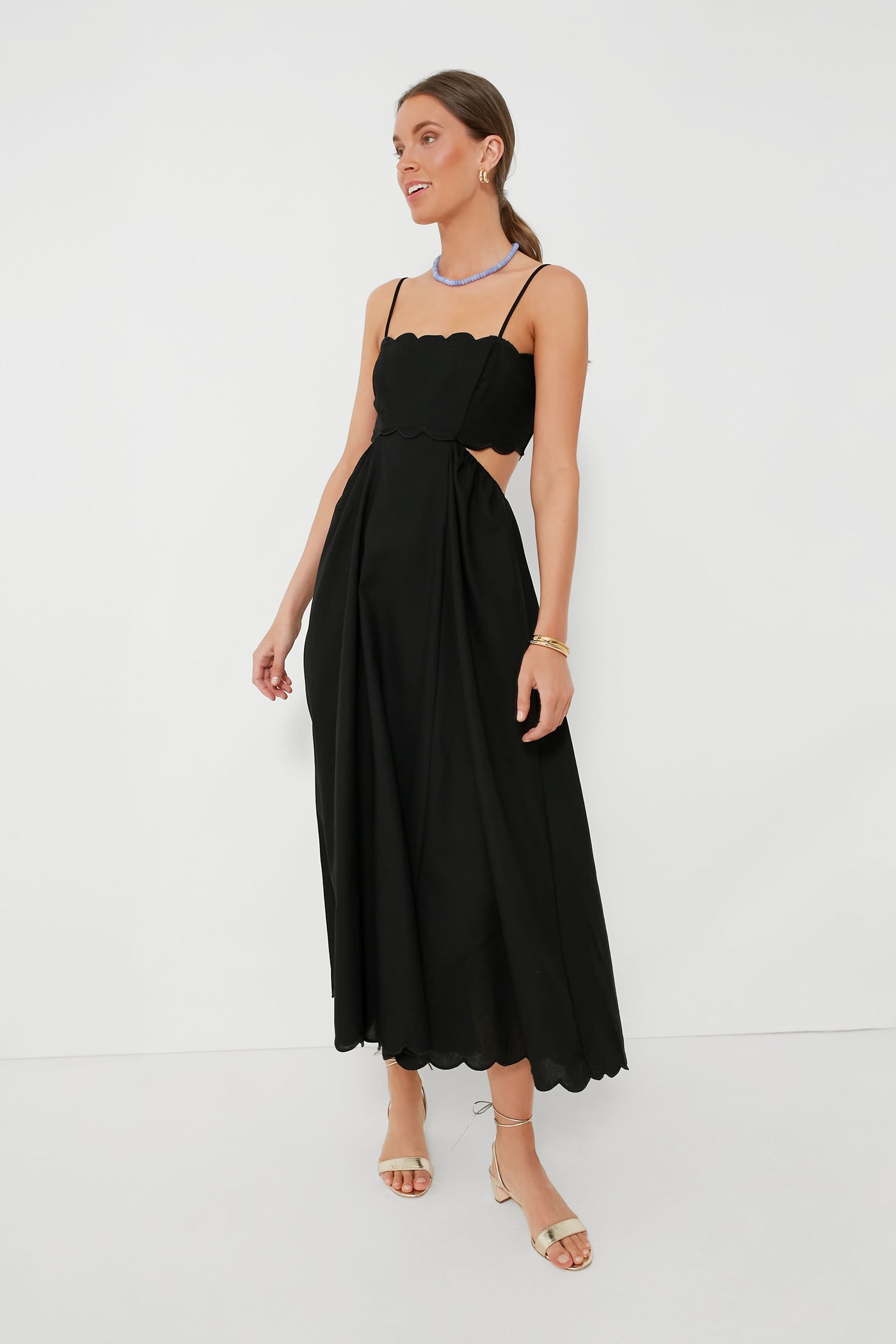 A woman wears a Moon River black dress with scallop trim, thin straps, open sides, minimal jewelry, and beige heeled sandals.
