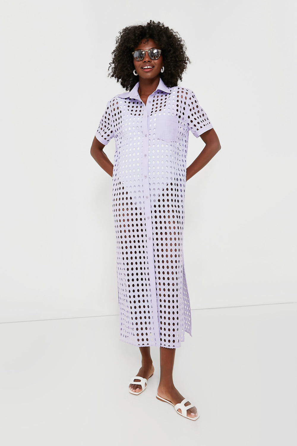 A woman in sunglasses, a lavender Wisteria Oxford Tunic shirt dress by Solid & Striped, and white sandals models chic beachwear.