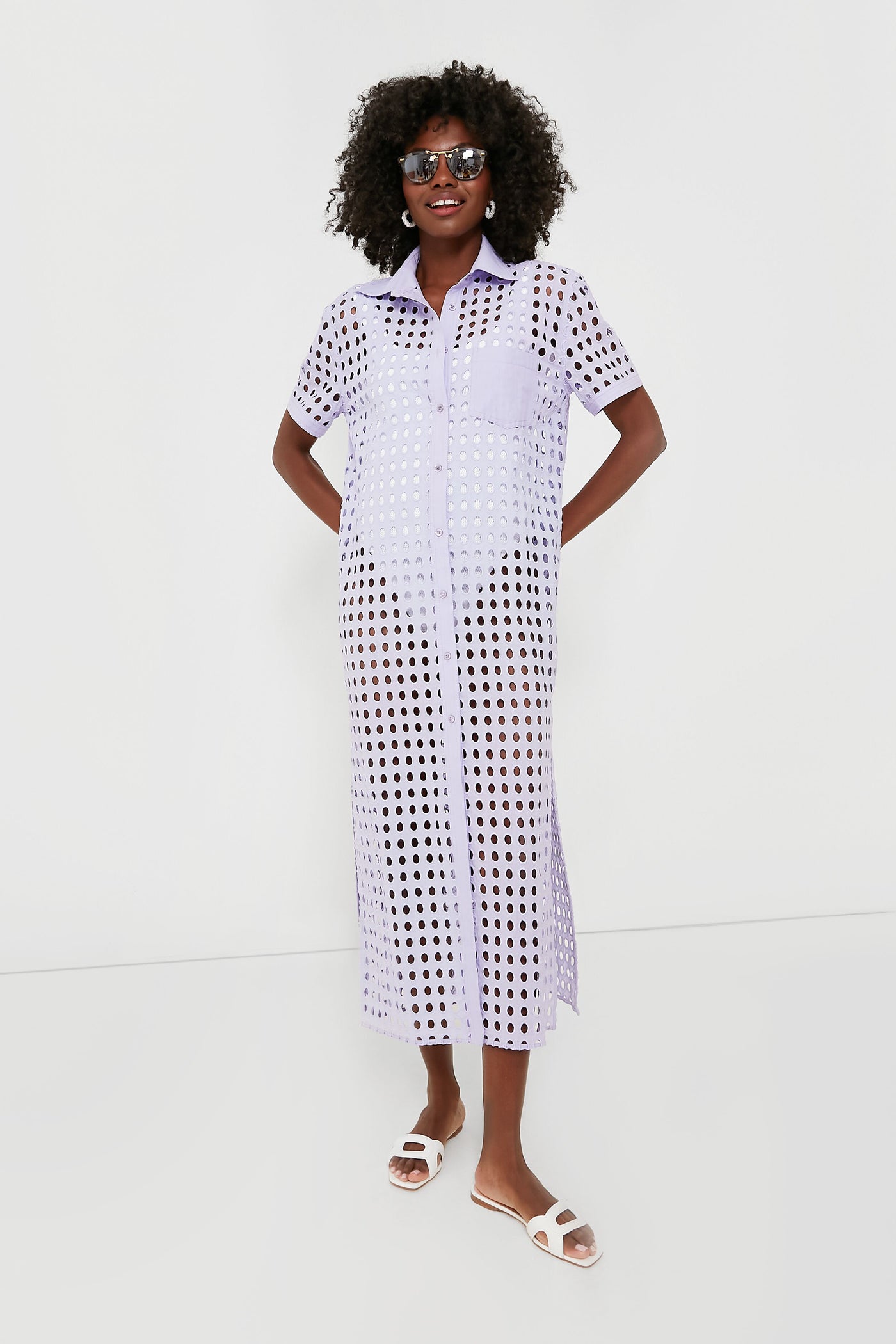 A woman in sunglasses, a lavender Wisteria Oxford Tunic shirt dress by Solid & Striped, and white sandals models chic beachwear.