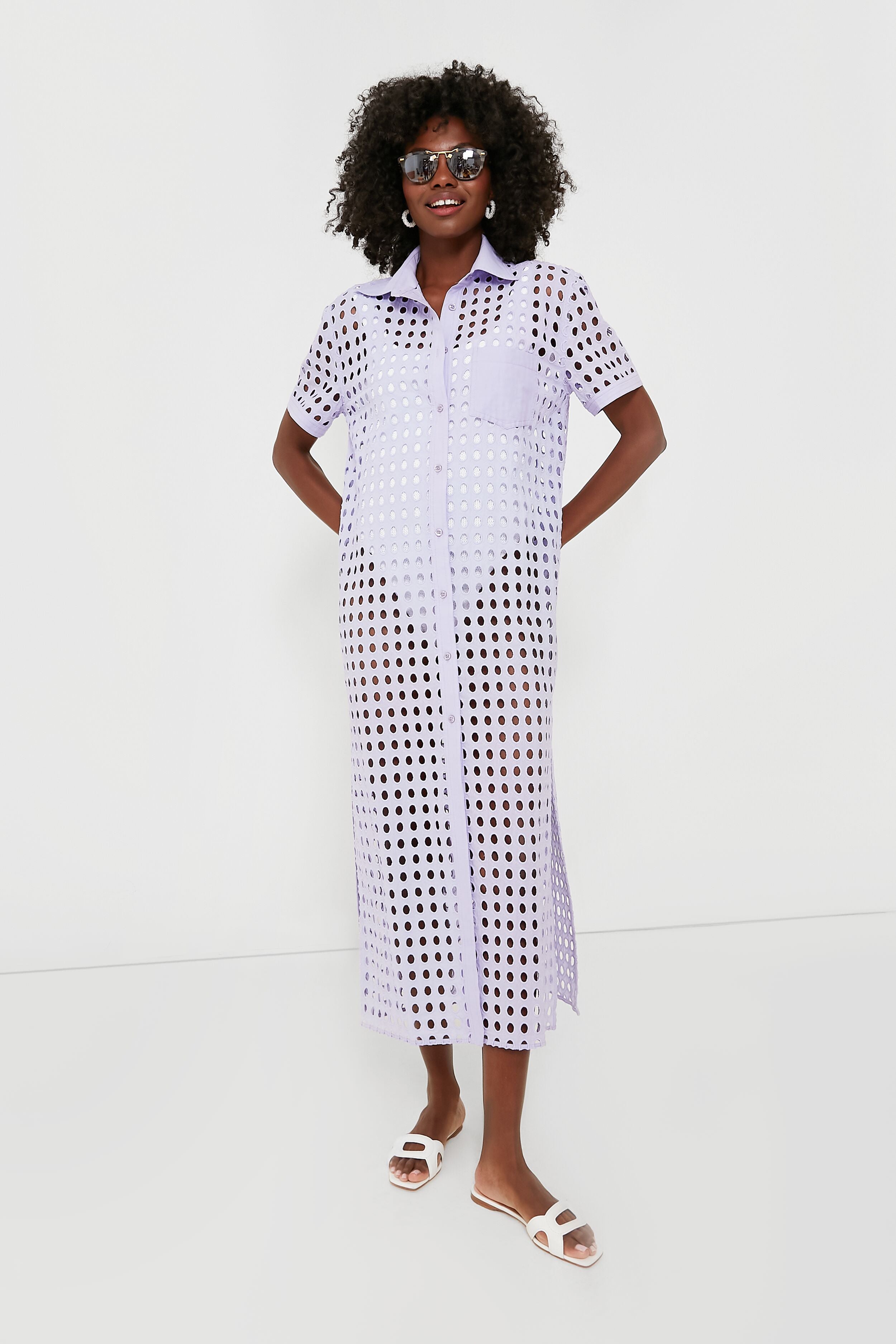 A woman in sunglasses, a lavender Wisteria Oxford Tunic shirt dress by Solid & Striped, and white sandals models chic beachwear.