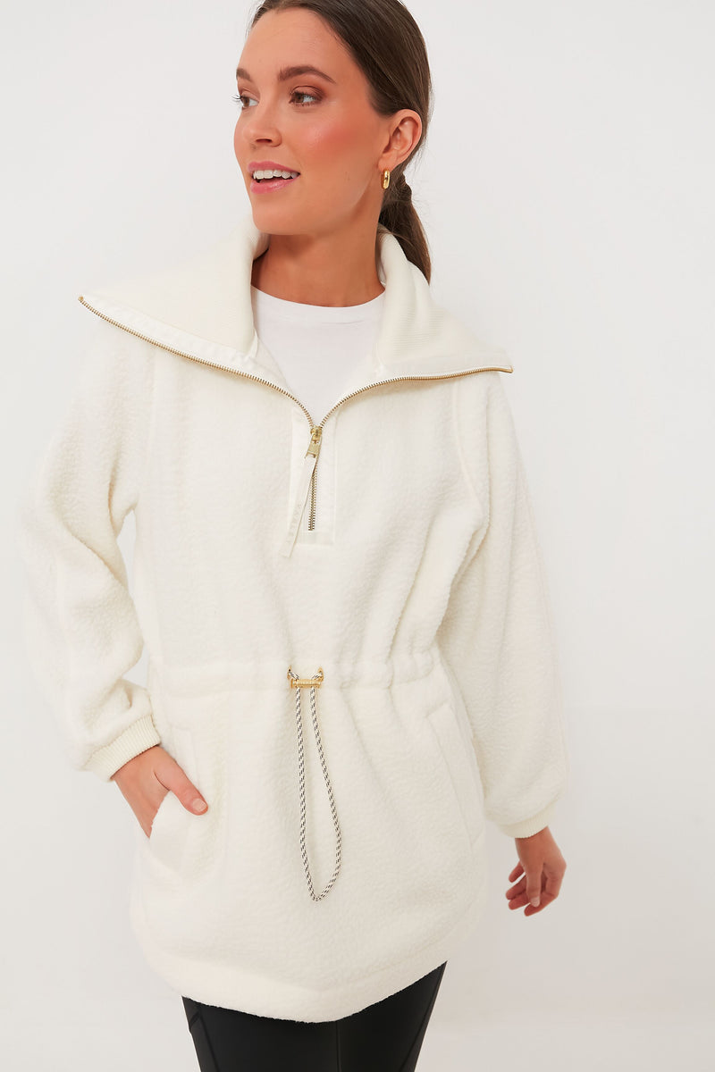A woman wears a white Varley Plush Borg Fleece zip-up jacket with a drawstring waist and black pants, exemplifying cozy alpine loungewear style.