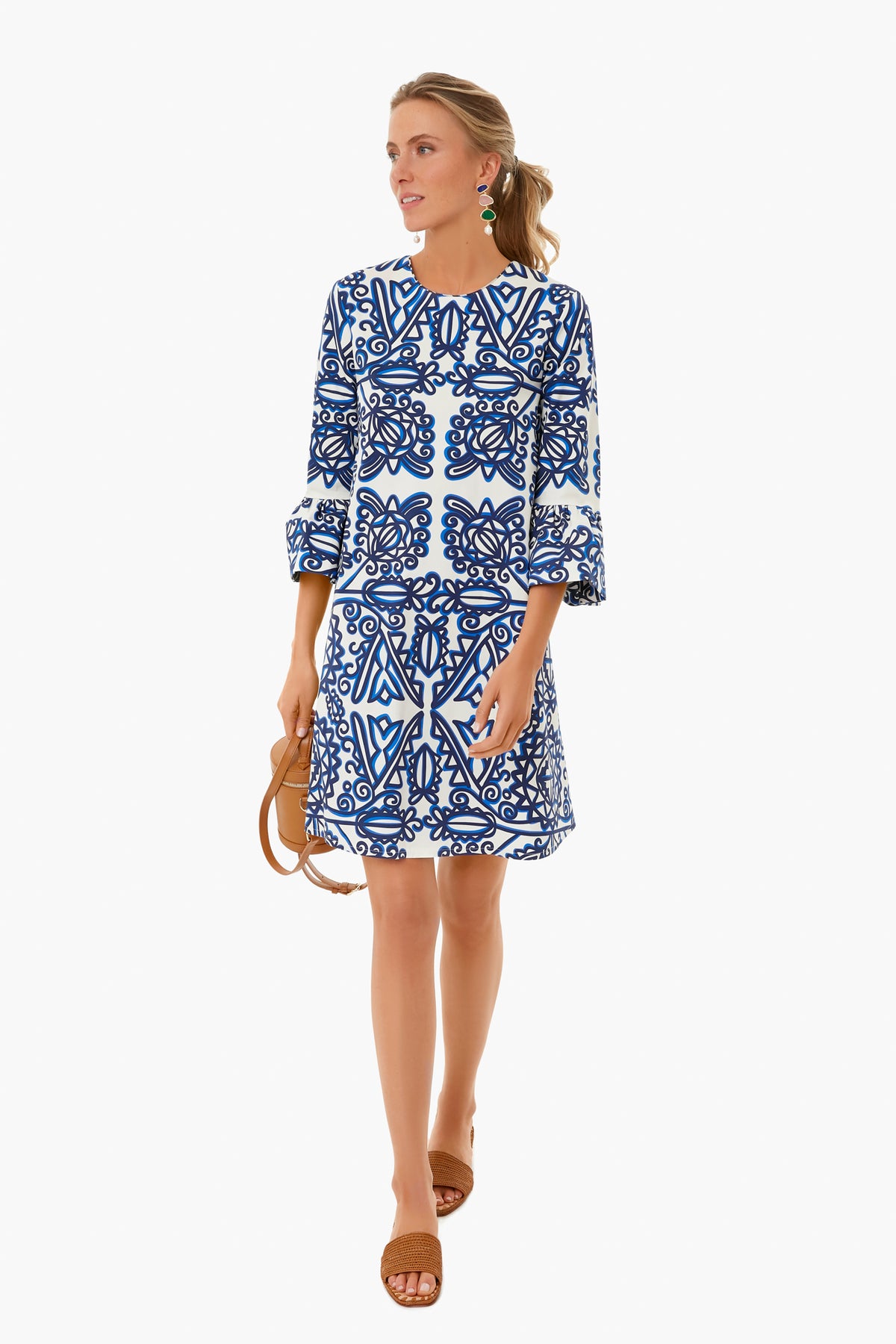 A woman showcases vintage-inspired Italian fashion in a blue and white La DoubleJ dress with three-quarter sleeves, holding tan sandals and wearing brown sandals.