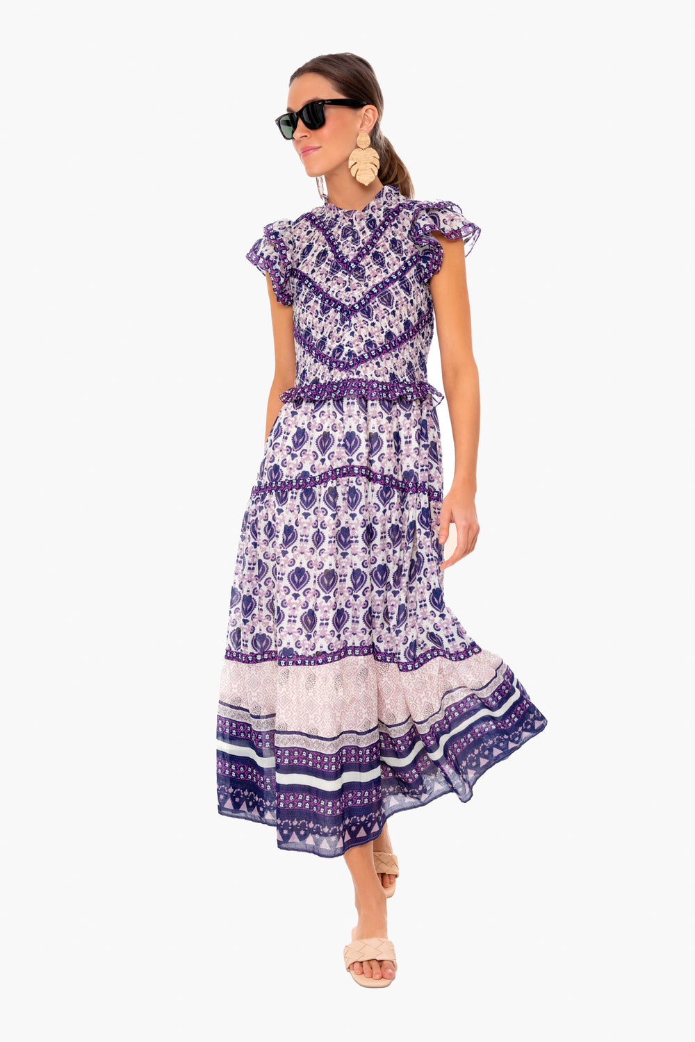 A woman in a Sea New York purple and white boho print midi dress with a smocked bodice, short sleeves, sunglasses, statement earrings, and sandals.