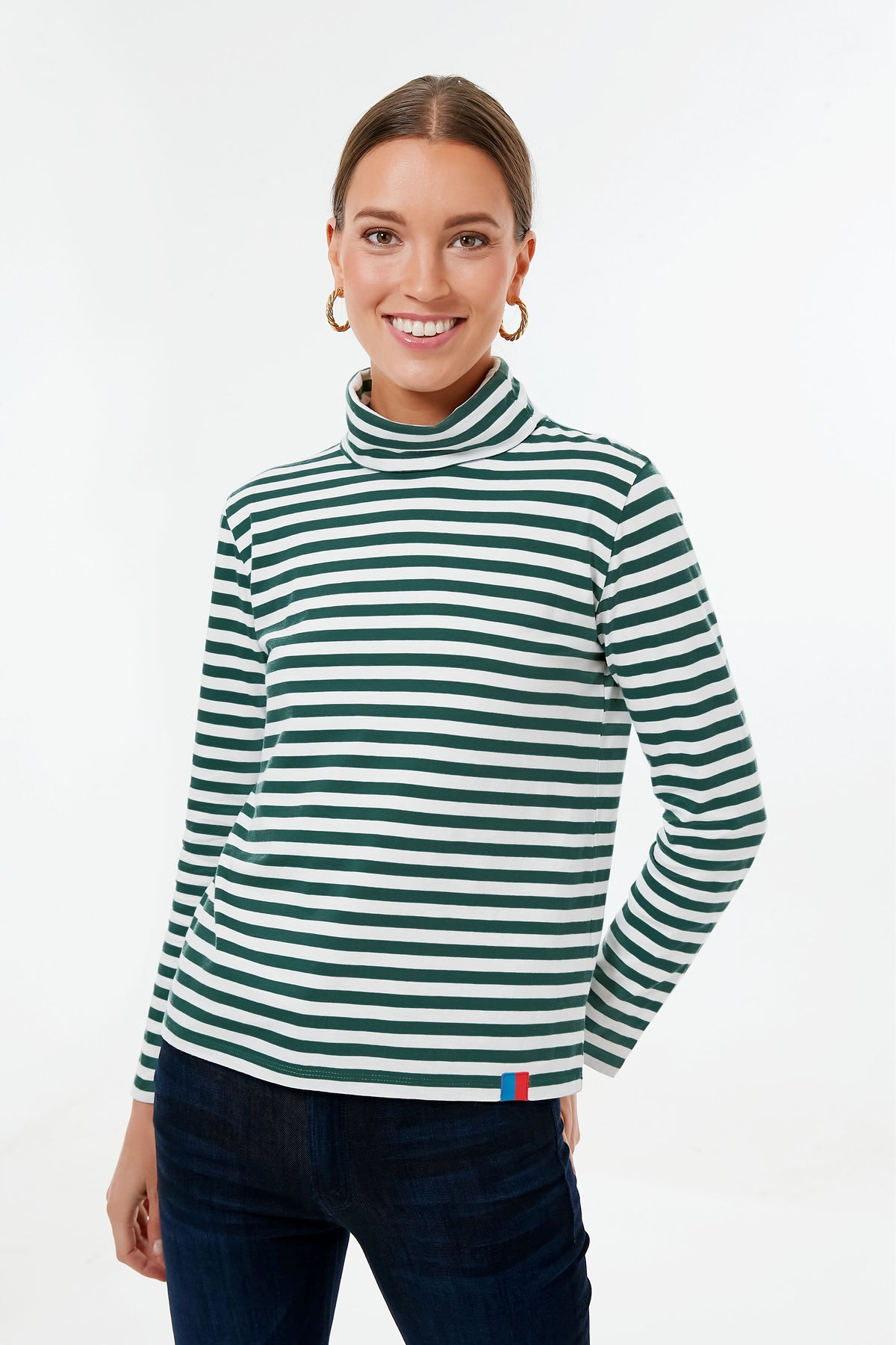A woman in a black and white stripe KULE turtleneck long-sleeve shirt and dark pants smiles at the camera, showcasing this perfect layering piece.